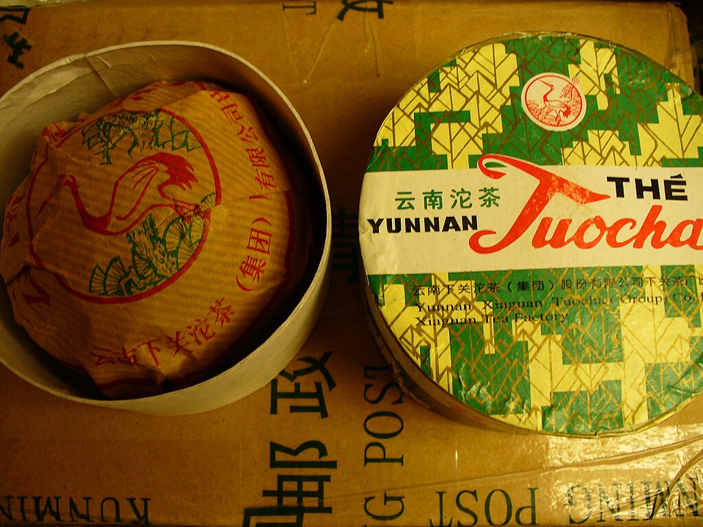 Emballage traditionnel pour une galette de thé pu-erh Xiaguan Tuocha du Yunnan, montrant une galette emballée dans une boîte ronde et une boîte étiquetée verte et jaune.