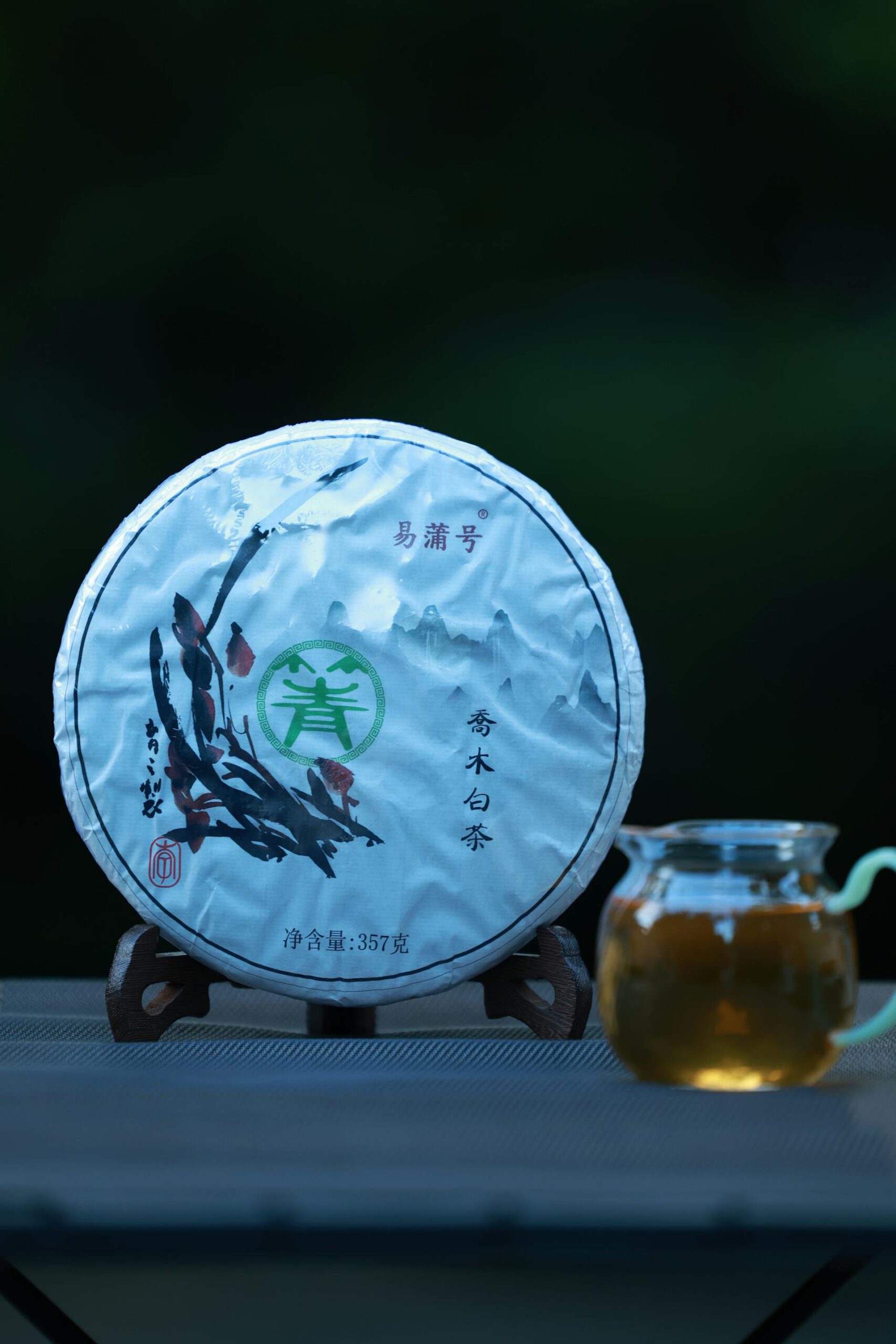 Une galette de thé pu-erh, emballée dans un papier traditionnel avec des caractères chinois, posée sur un support en bois à côté d'une théière en verre contenant du thé infusé.