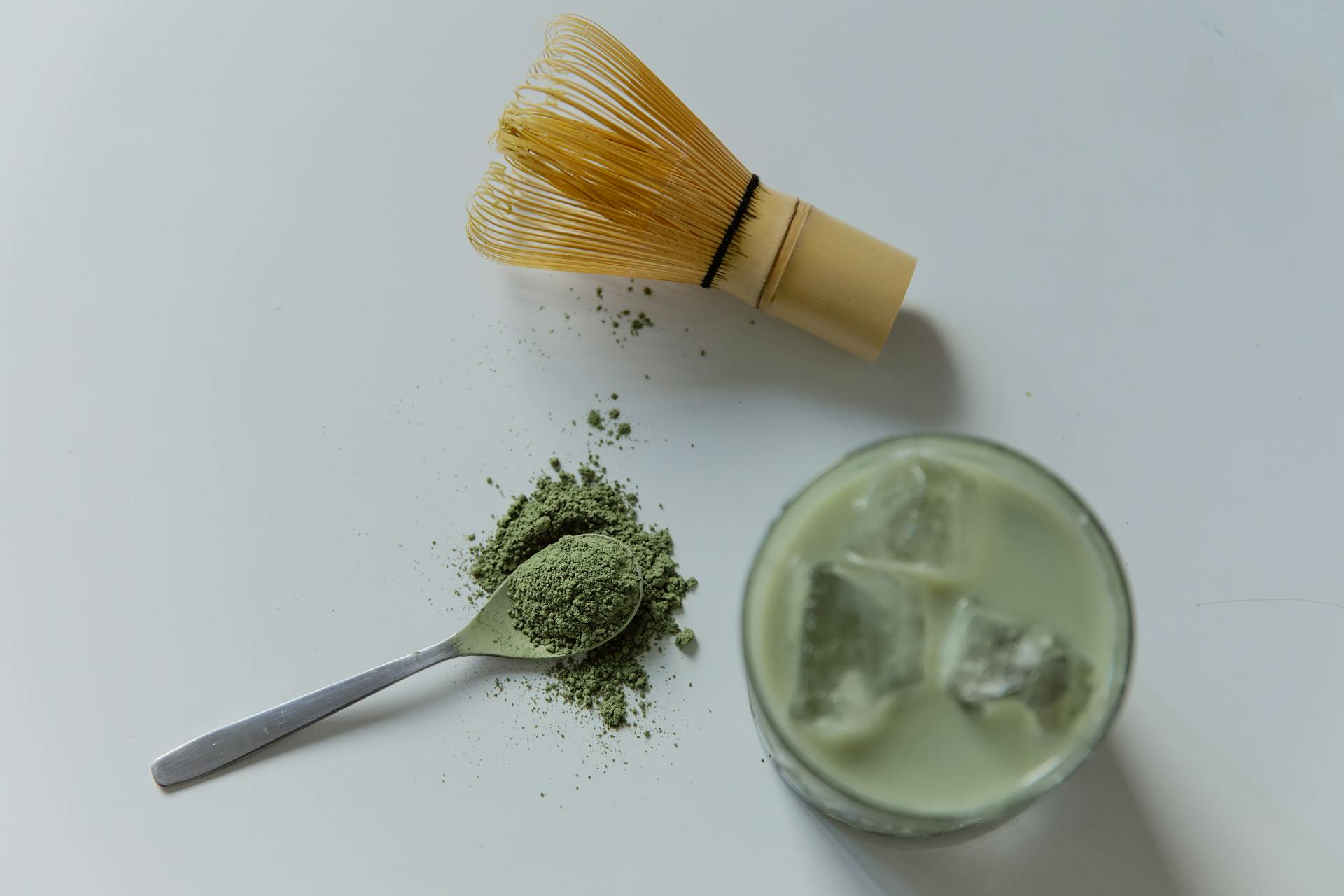 Vue de dessus des ingrédients clés pour un latte au matcha chinois glacé : un fouet en bambou, une cuillère de poudre de matcha verte et un verre de la boisson froide finie avec des glaçons.