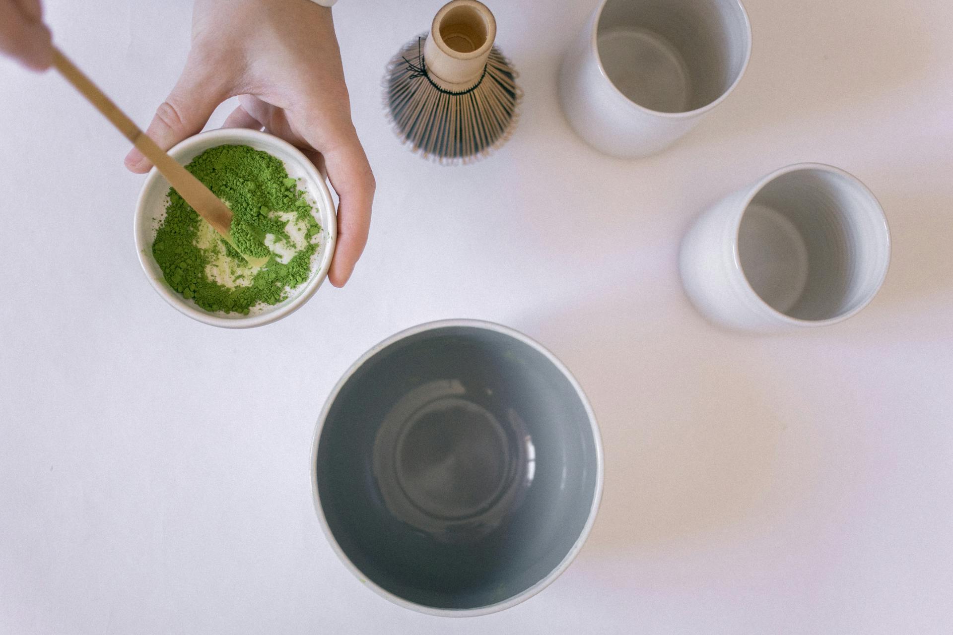 Vue de dessus d'une personne mélangeant de la poudre de thé vert matcha dans un bol blanc avec une cuillère en bois, montrant la première étape d'une préparation traditionnelle du matcha.