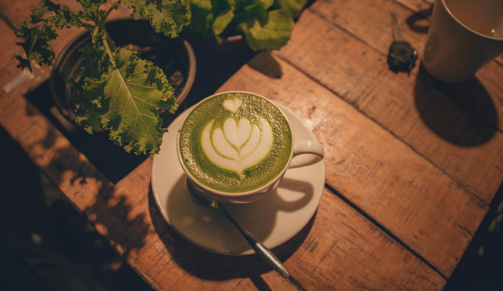 Une photo esthétique d'un latte au matcha vert avec un dessin de cœur en latte art, servi sur une table en bois avec une cuillère et une plante, idéal pour un café ou un blog de style de vie.