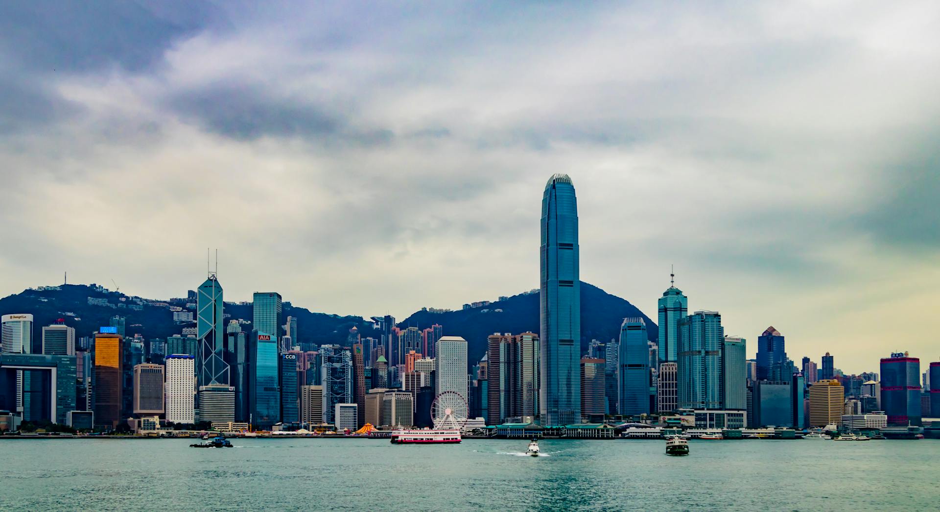Skyline de Hong Kong avec ses gratte-ciel et le port Victoria au premier plan sous un ciel nuageux.