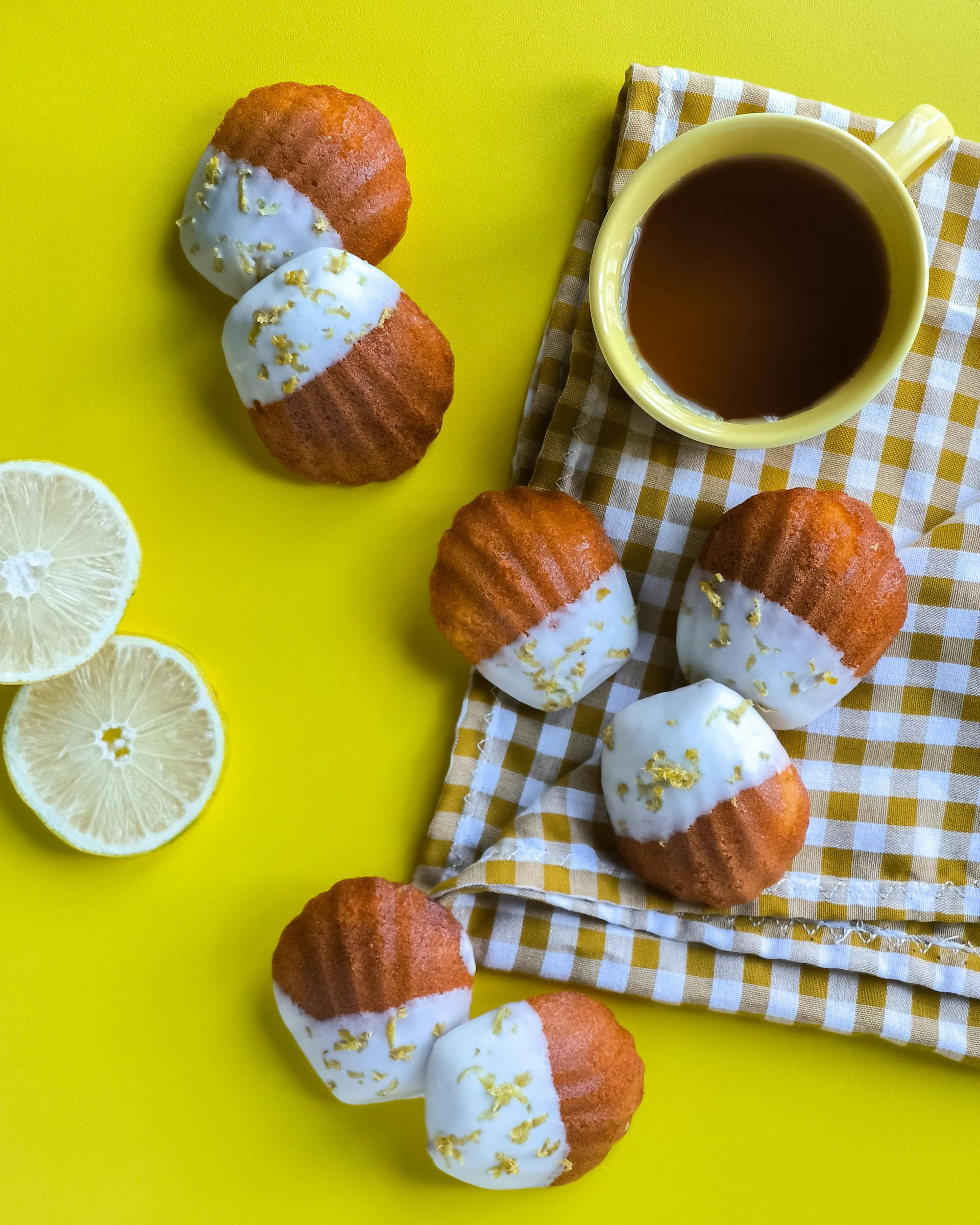Un assortiment festif de madeleines au citron, certaines trempées dans du chocolat blanc et des paillettes d'or comestibles, à côté d'une tasse de thé sur un fond jaune, pour un article sur les accords thé et dessert.