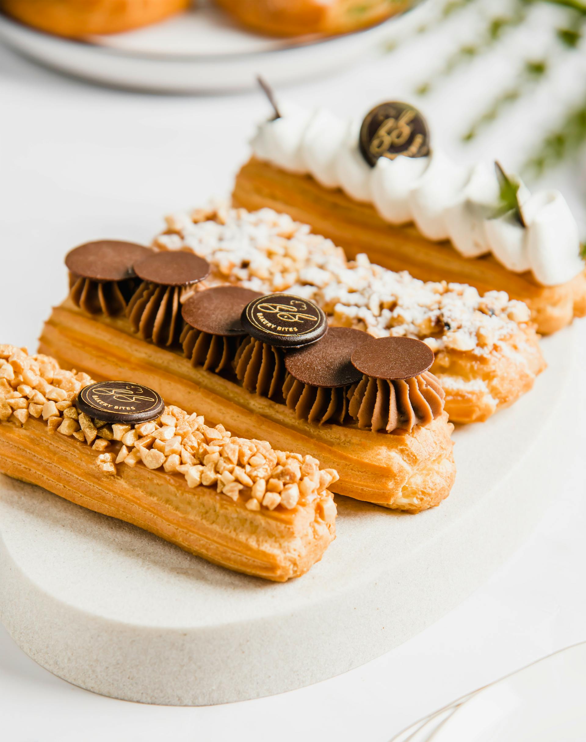 Trois éclairs élégants et délicieux avec différentes garnitures, notamment du chocolat, du caramel et des noix hachées, présentés sur une assiette de service blanche pour une recette de dessert ou une pâtisserie.