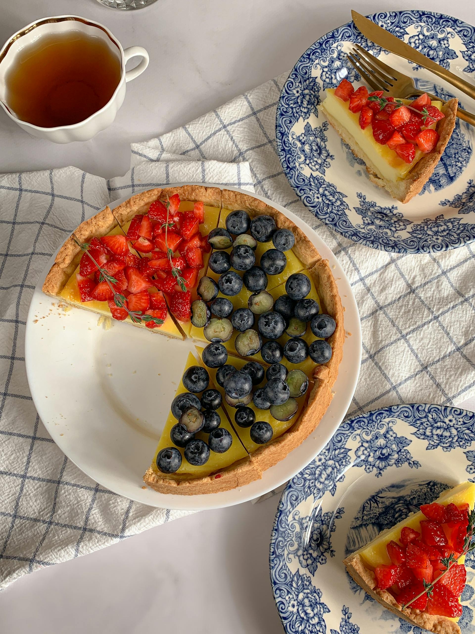 Une tarte aux fruits joliment décorée et coupée en morceaux, avec une garniture jaune vif surmontée de fraises et de myrtilles fraîches, servie avec du thé pour un article de blog sur les desserts.