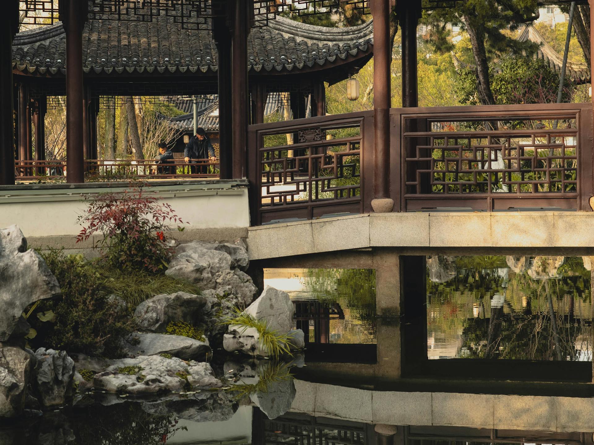 Un jardin chinois paisible à Suzhou, en Chine, avec un pavillon traditionnel en bois et un pont de pierre se reflétant dans un étang calme, illustrant l'architecture paysagère classique d'Asie de l'Est.