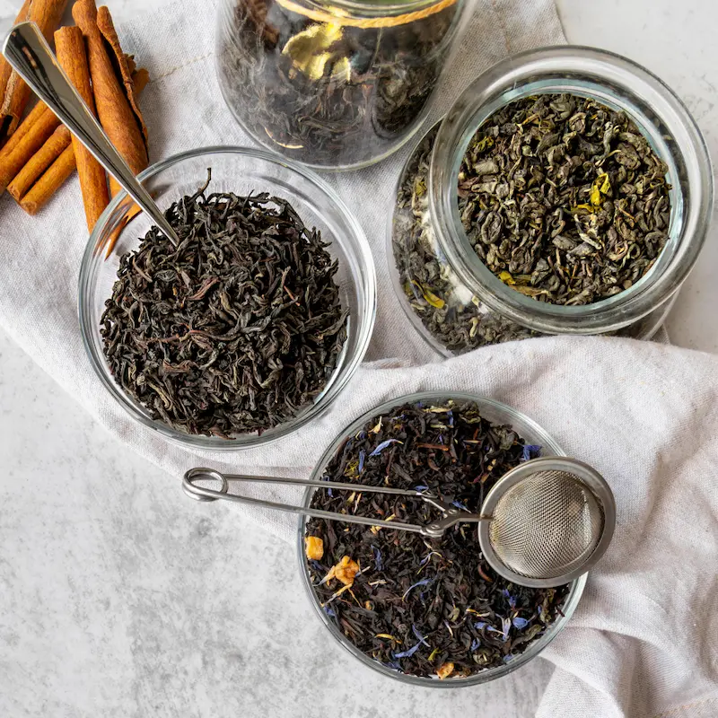 Conservation du thé : Différents types de thé noir, thé vert et mélanges floraux séchés stockés dans des bols et bocaux en verre pour le stockage. Une pince-infuseur et de la cannelle montrent la préparation d'une infusion fraîche.