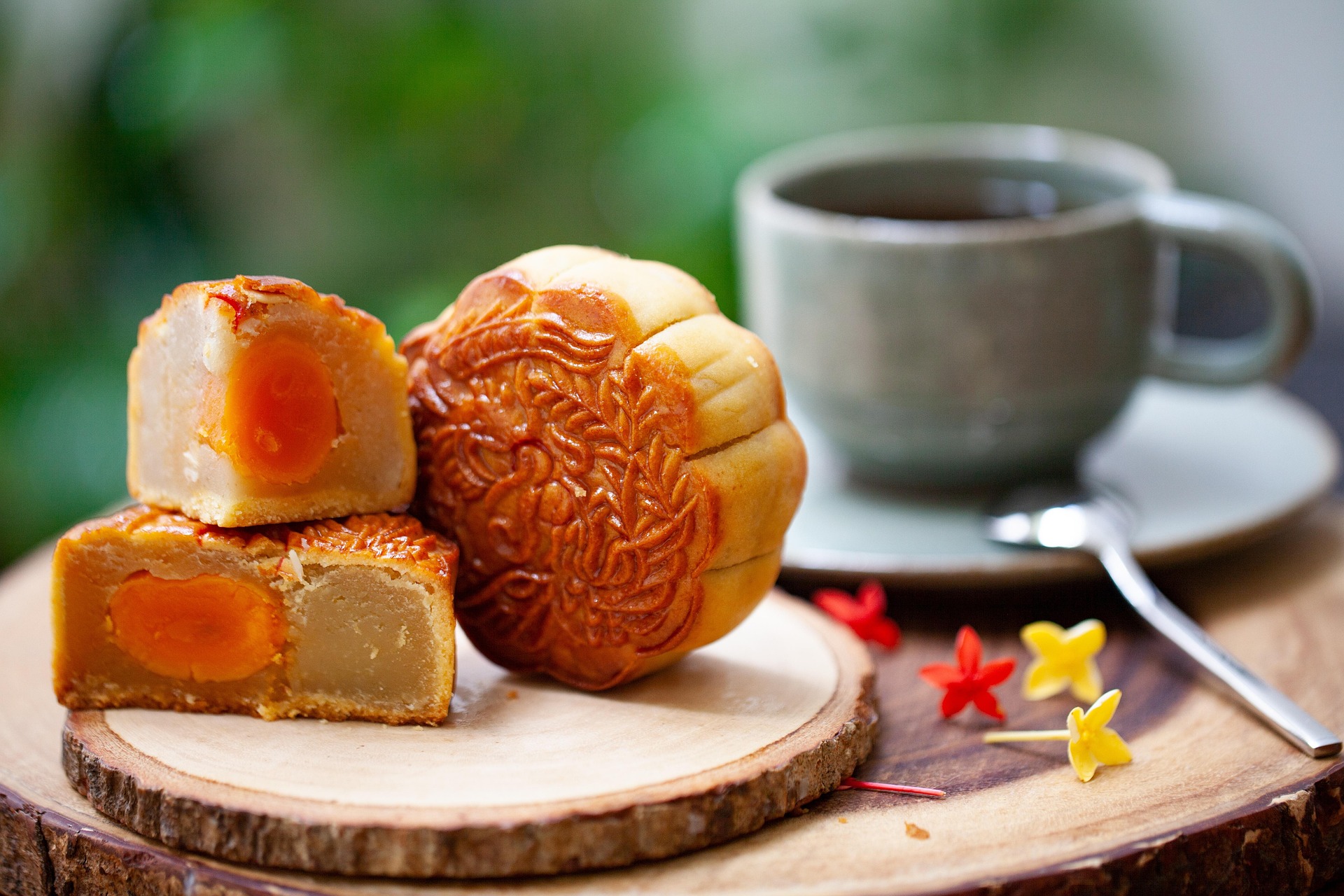 Deux moitiés d'un Mooncake traditionnel aux jaunes d'œuf salés, posées sur une souche en bois. En arrière-plan flou, une tasse de thé est prête. L'image illustre le parfait moment de dégustation et les accords thé mooncake pour la Fête de la Mi-Automne.