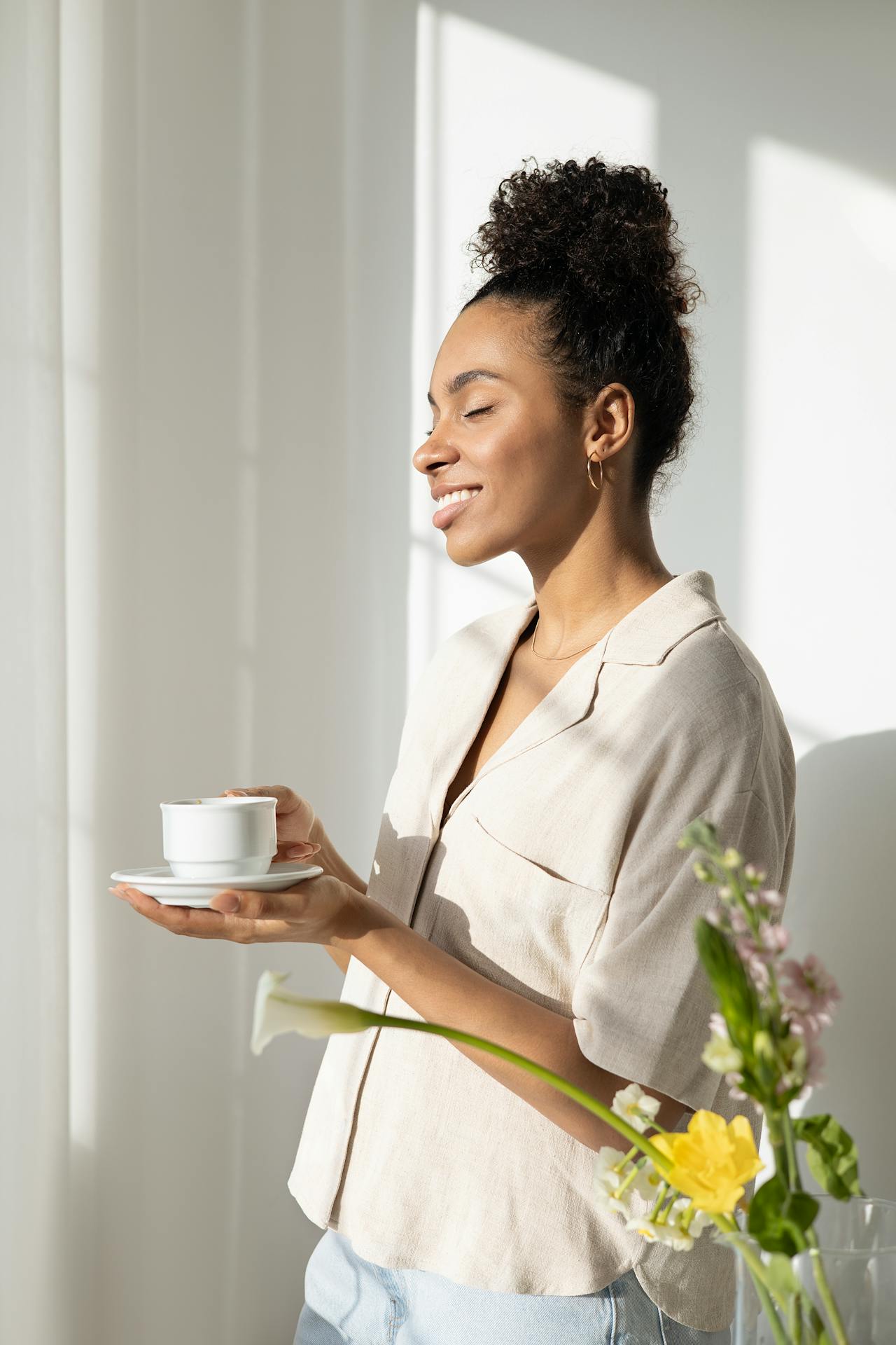 Femme souriante, les yeux fermés, dégustant une tasse de thé fumante dans un intérieur lumineux. Moment de détente, sérénité et bien-être matinal.