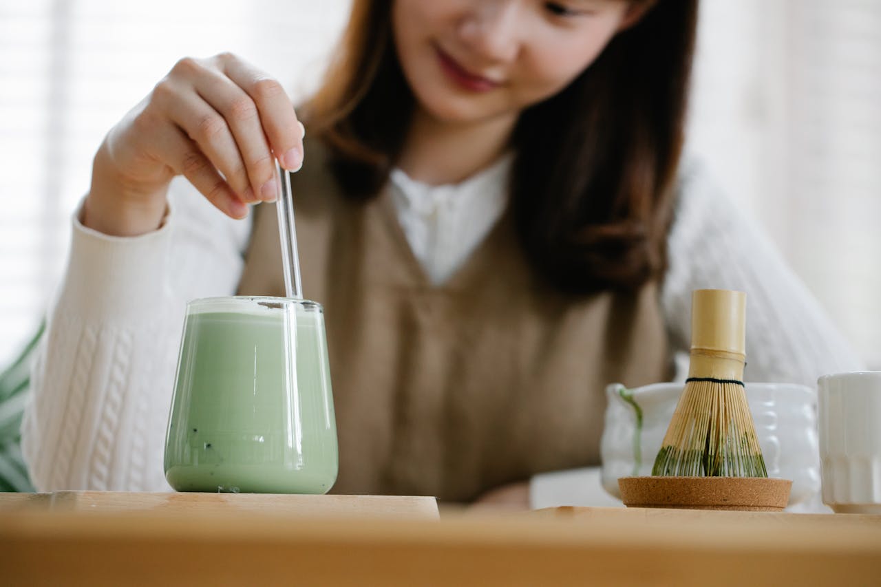 Jeune femme mélangeant un matcha latte glacé (vert pastel) dans un verre transparent. Fouet en bambou (chasen) et tasse de thé japonais en arrière-plan.