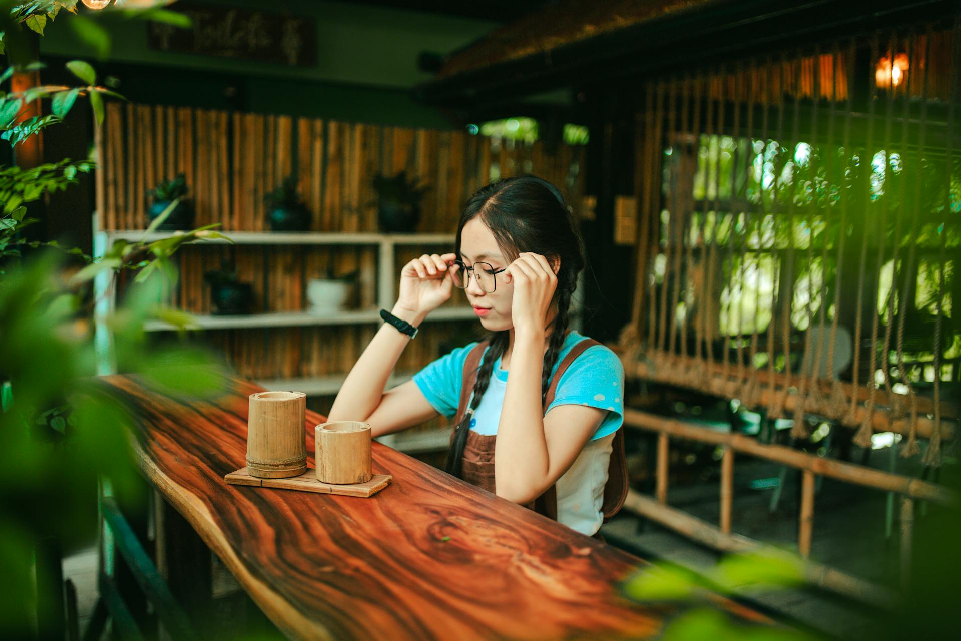 Jeune femme ajustant ses lunettes, assise seule à un bar en bois exotique dans une ambiance de salon de thé asiatique. Moments de réflexion et de clarté mentale avec une tasse de thé chinois traditionnel.
