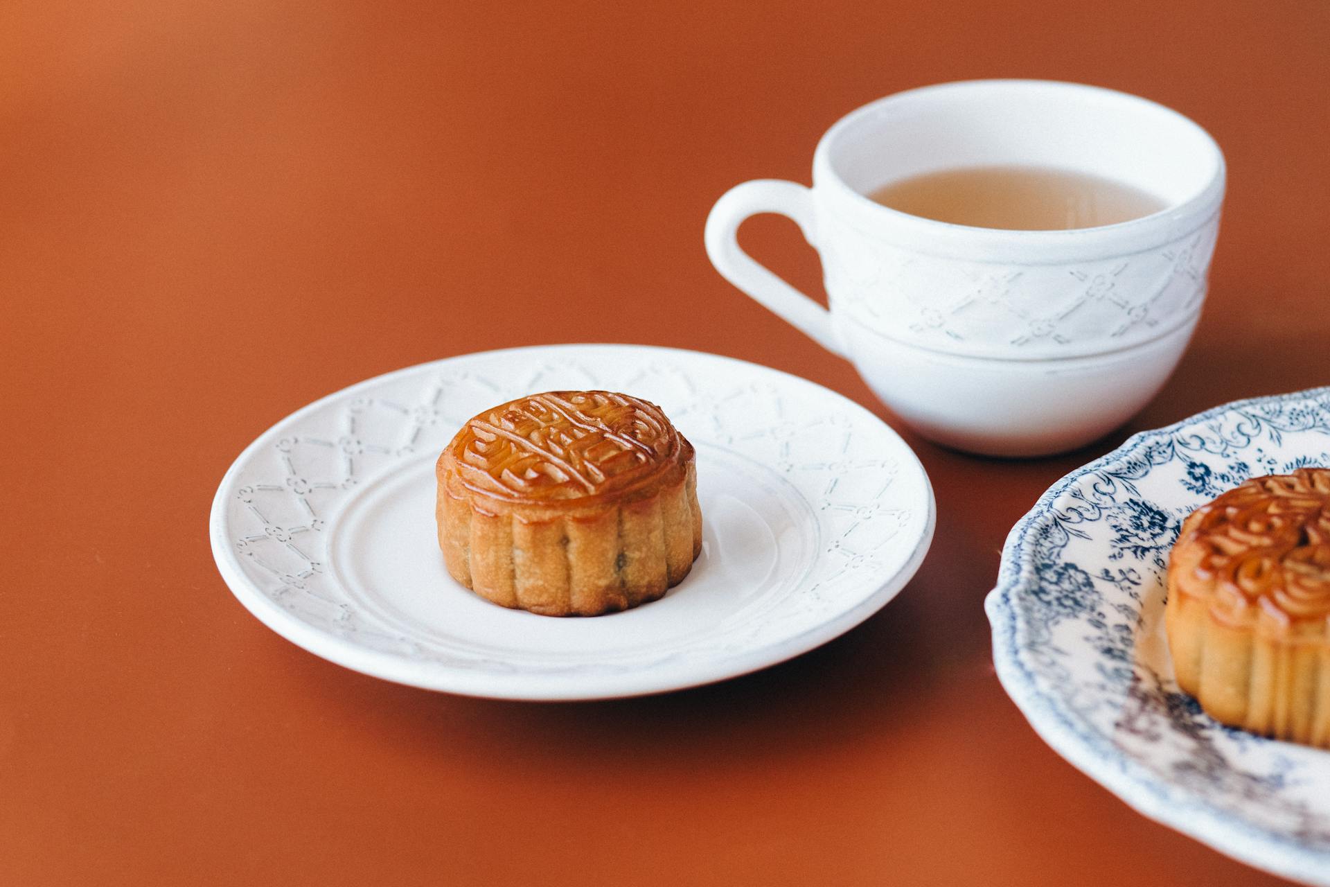 Un Mooncake doré et rond, avec des motifs traditionnels, posé sur une petite assiette en céramique blanche. À côté, une tasse de thé vert est prête. La scène illustre un moment de dégustation calme et la réussite des accords thé mooncake pour la Fête de la Mi-Automne.
