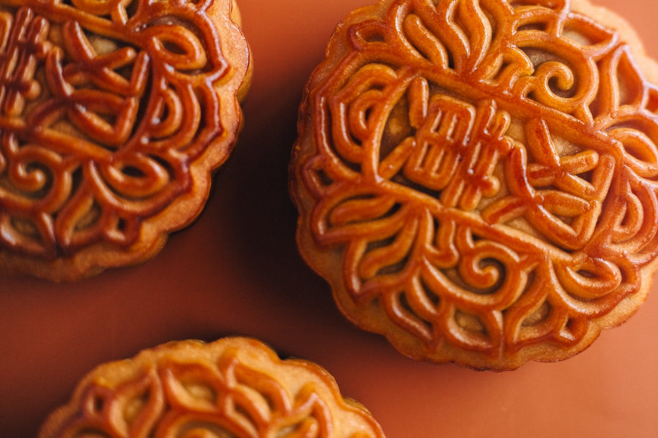 Gros plan détaillé sur trois Mooncakes dorés traditionnels posés sur un fond de couleur terre cuite. Les motifs chinois en relief et la texture brillante des gâteaux de lune sont visibles. Cette image symbolise le début de la Fête de la Mi-Automne et la recherche des accords thé-mooncake parfaits.