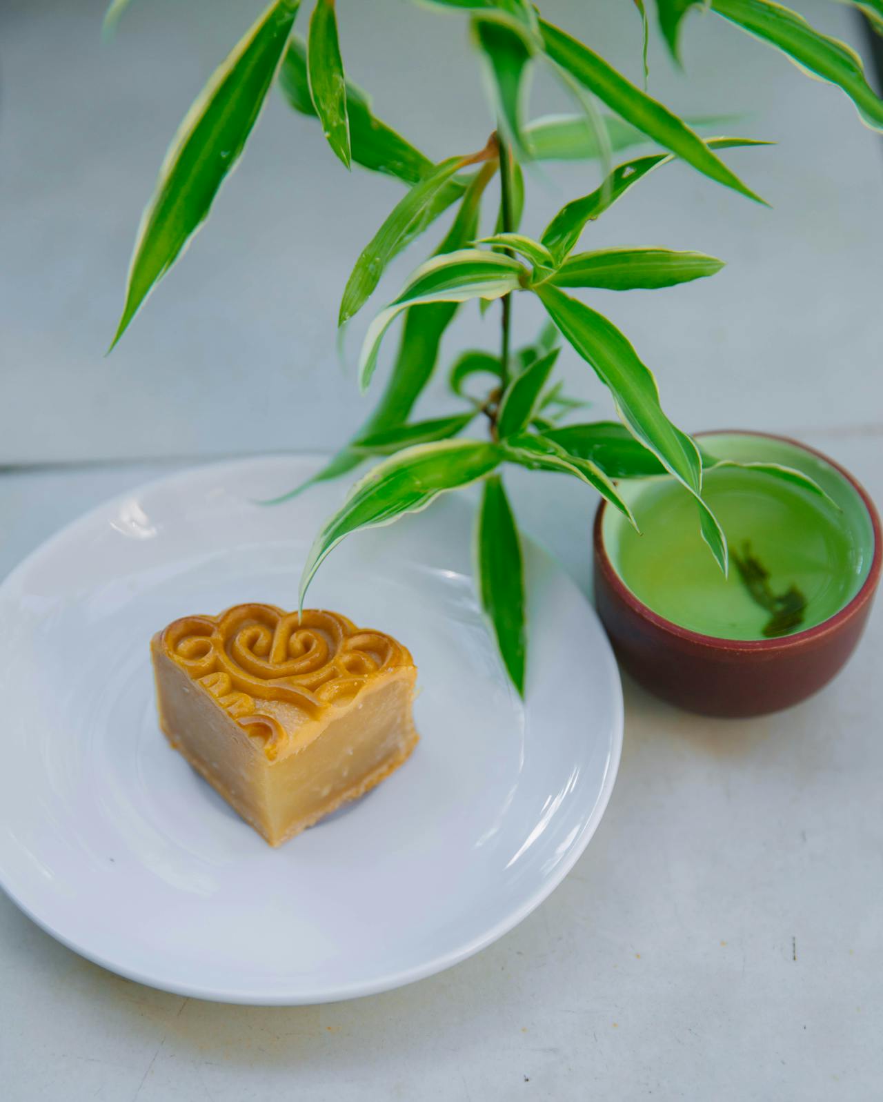 Mooncake moderne en forme de cœur sur une assiette blanche, accompagné d'une tasse de thé vert clair. Une plante verte et fraîche en arrière-plan. L'image illustre un moment zen et de délicieux accords thé mooncake.