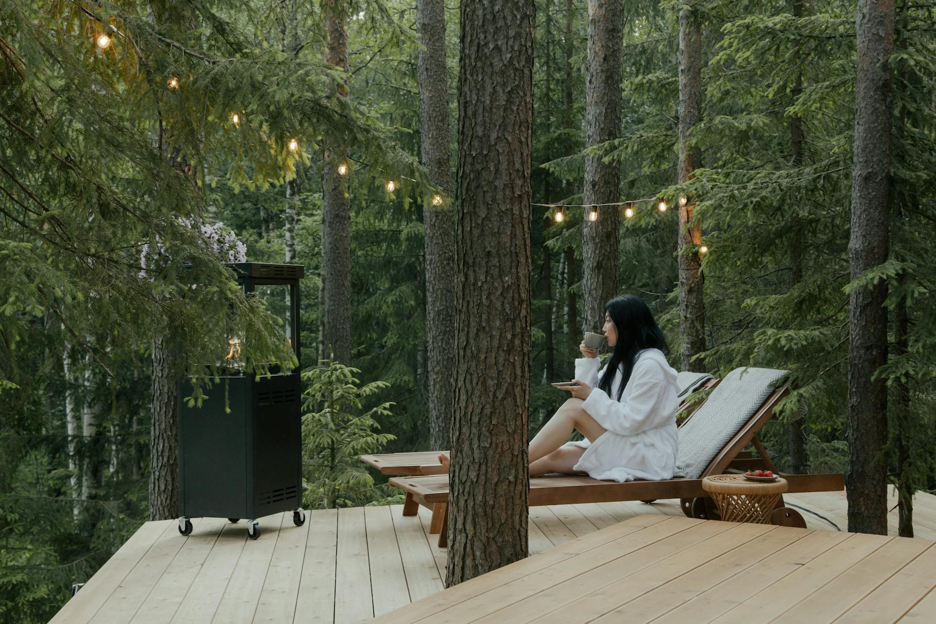 Femme en peignoir blanc dégustant un thé chaud sur une terrasse en bois aménagée au milieu d'une forêt de pins. Guirlande lumineuse et chauffage extérieur créant une ambiance hygge et détente.