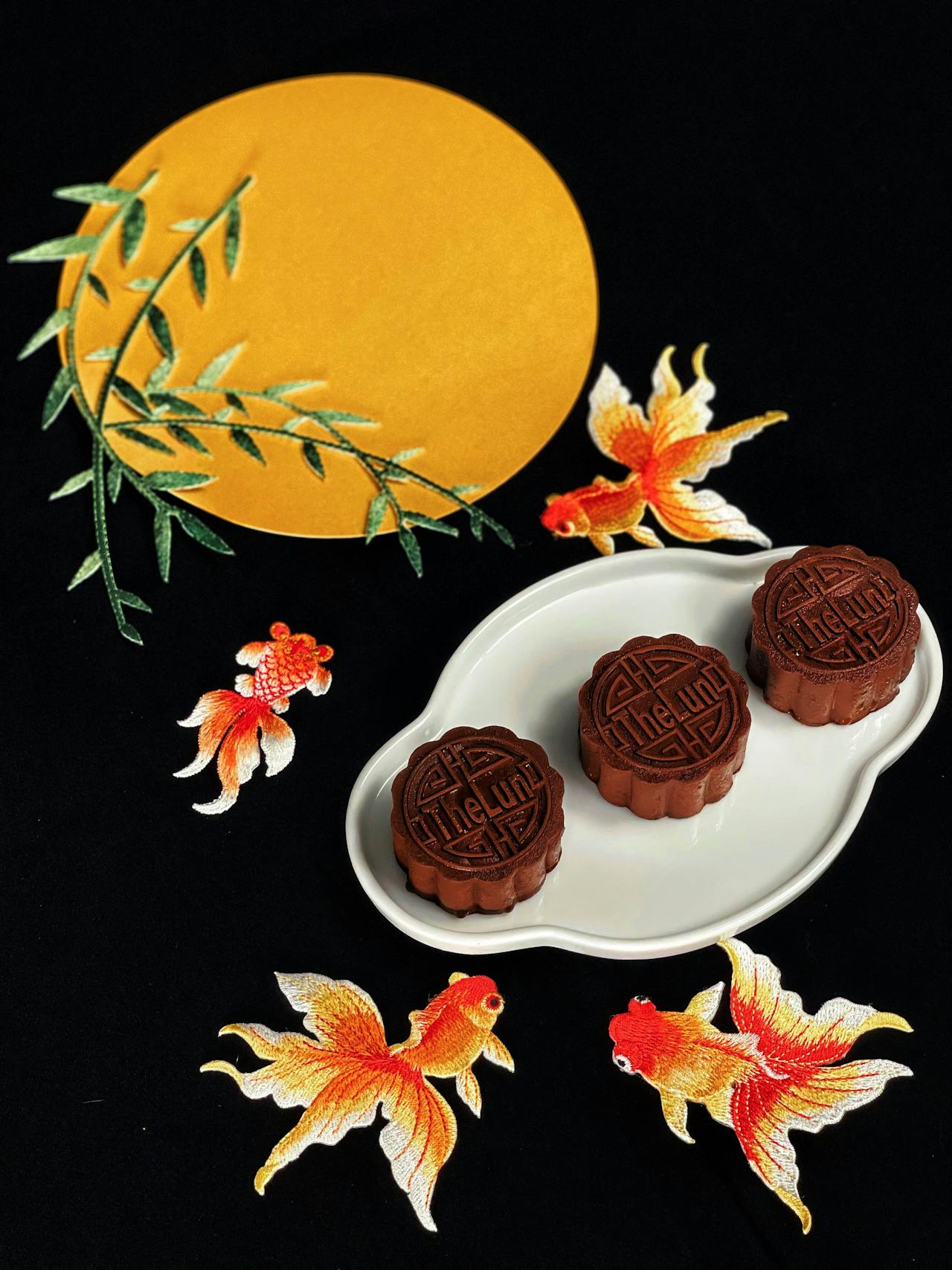 Composition créative pour la Fête de la Mi-Automne. Trois Mooncakes modernes au chocolat sont présentés sur une assiette blanche, entourés de broderies de poissons rouges et d'une lune jaune. Cette image illustre l'un des accords thé mooncake modernes pour le thé noir.