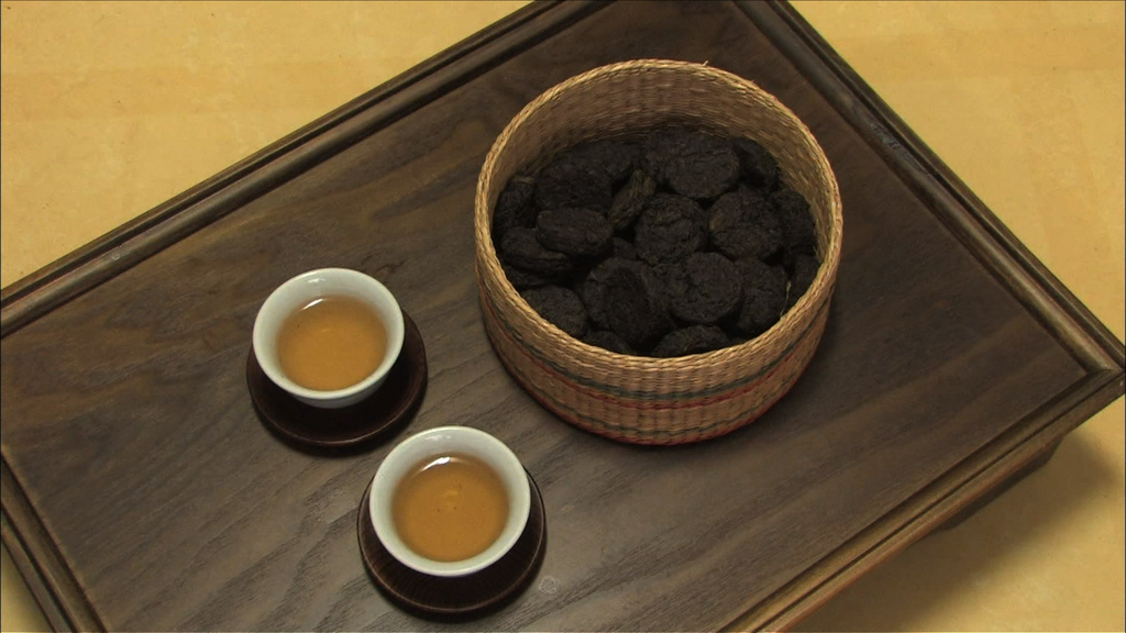 Vue de dessus d'un panier en osier rempli de petites galettes ou nids de thé compressé sombre de forme ronde (thé Pu-Erh individuel). Deux petites tasses de thé infusé de couleur ambrée sont posées sur des soucoupes sur un plateau en bois foncé.