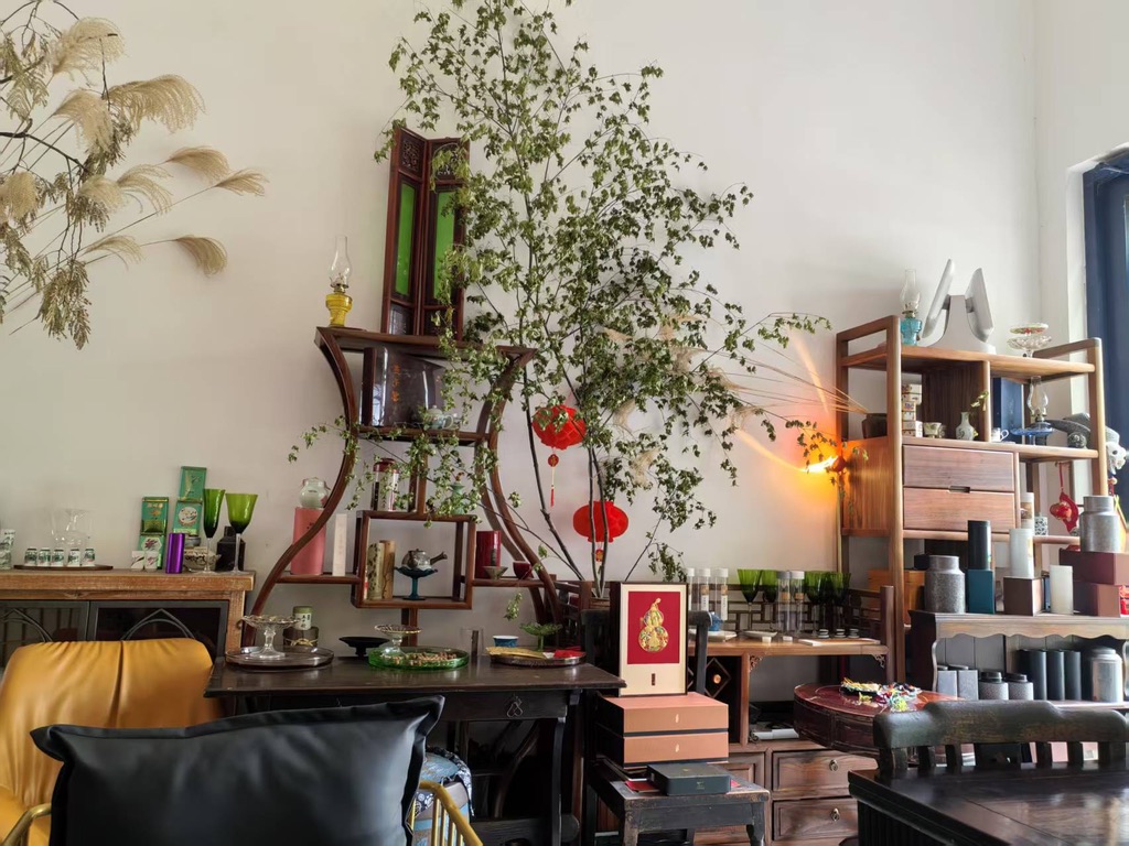 Intérieur d'un salon de thé chinois décoré dans un style traditionnel. La pièce est remplie d'étagères en bois sombre contenant diverses boîtes de thé chinois, de tasses, et d'accessoires de cérémonie. Un grand arrangement végétal se trouve au centre, et des boîtes cadeaux (possiblement du Pu-Erh) et des lanternes sont visibles.