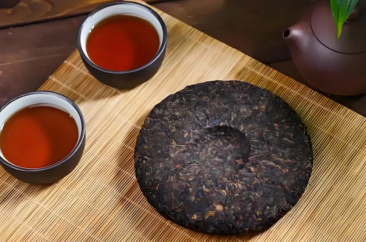 Galette de thé Pu-Erh fermenté de forme ronde, posée sur un tapis en bambou. Deux tasses noires de thé infusé et une théière en terre cuite sont visibles sur la droite. Image illustrant la préparation traditionnelle du thé Pu-Erh.