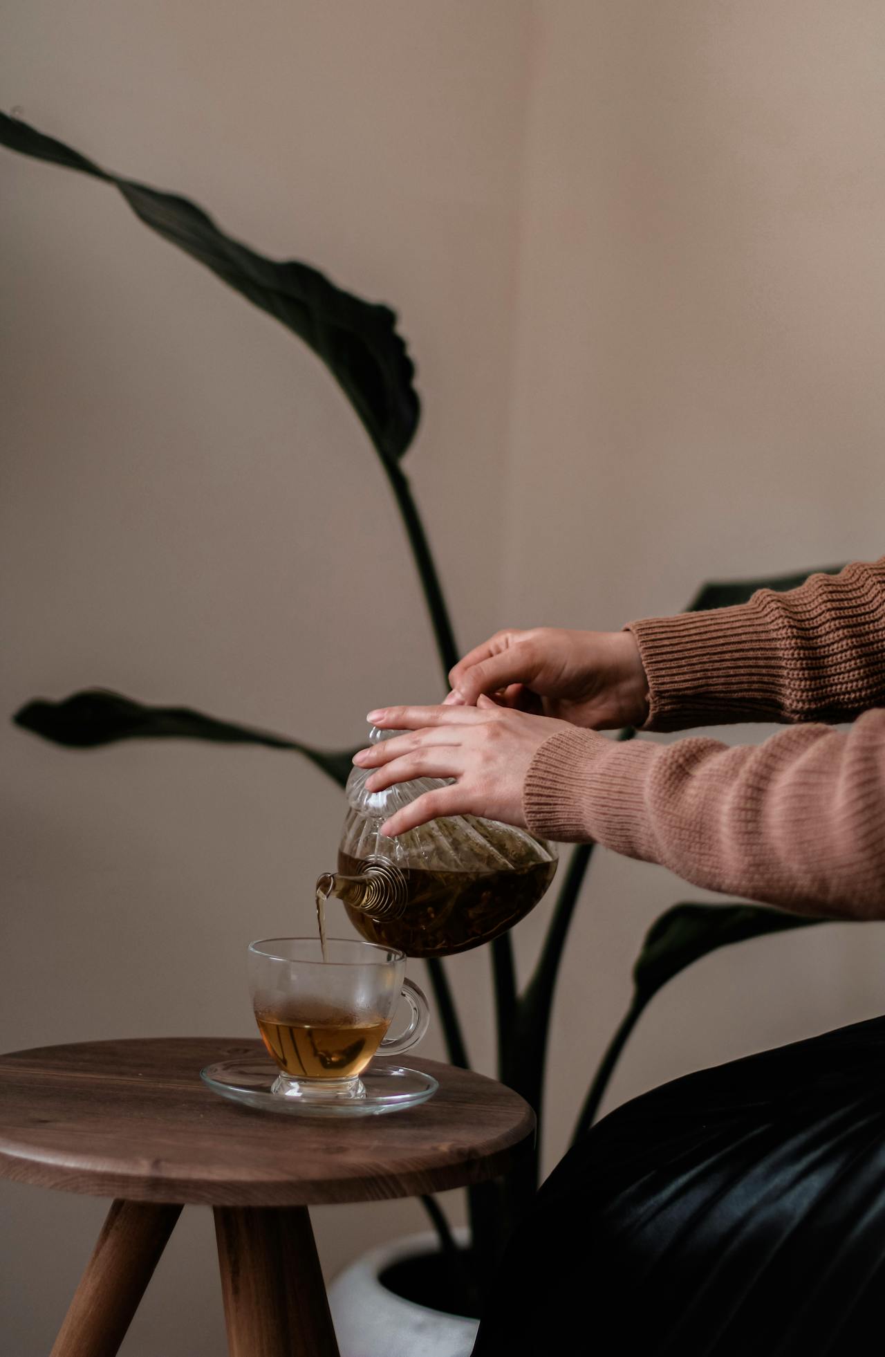 Gros plan sur deux mains versant du thé chaud (idéalement du thé Pu-Erh) d'une théière en verre dans une tasse transparente posée sur une petite table en bois. Cette image de rituel et de calme symbolise la santé et l'apaisement de la digestion.