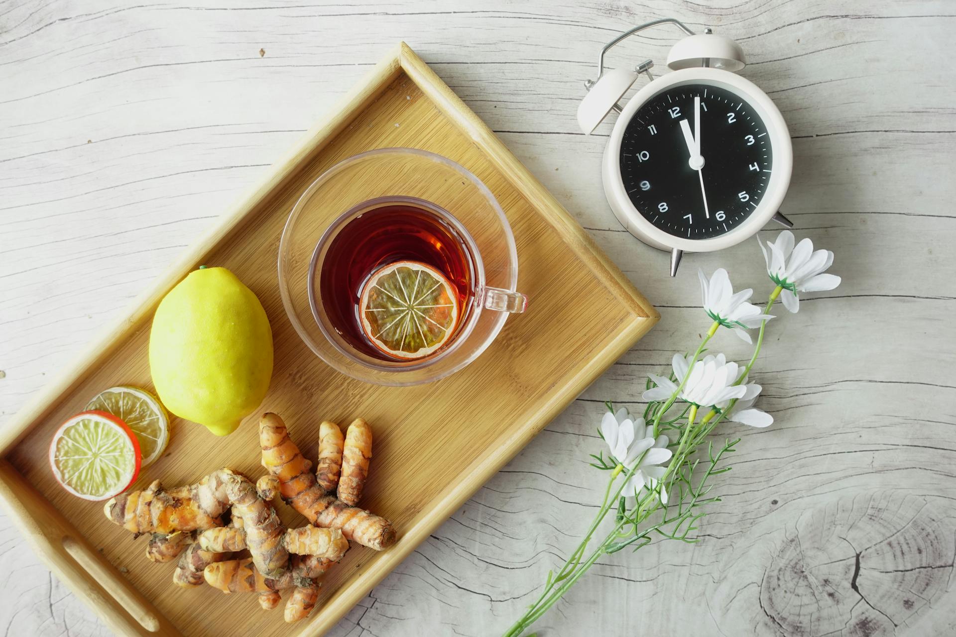 Plateau en bois avec tasse d'infusion chinoise au citron et gingembre, racines de curcuma, citron frais, horloge et fleurs blanches, symbolisant un rituel bien-être pour la santé.