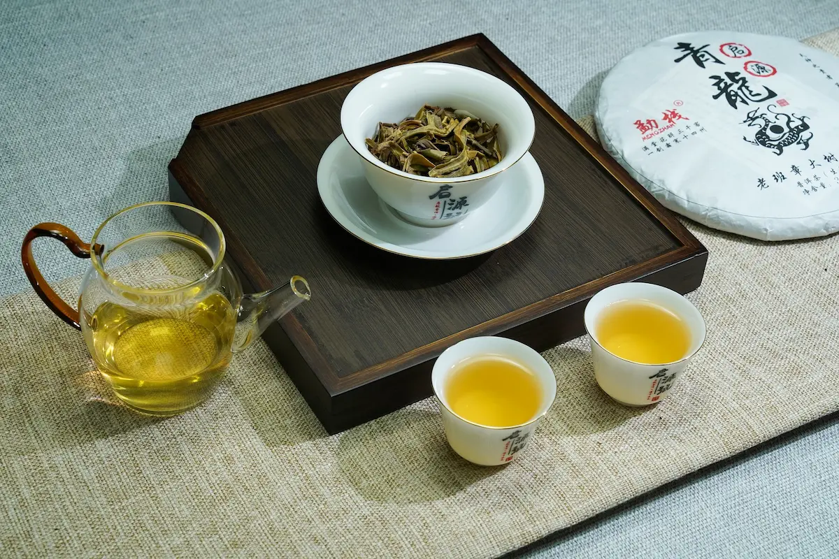 Ensemble de service de thé montrant une galette de Pu-Erh blanche et noire à droite, un bol de feuilles de thé Pu-Erh sèches, et deux petites tasses remplies de thé infusé de couleur or. Une petite théière en verre contient également le thé.
