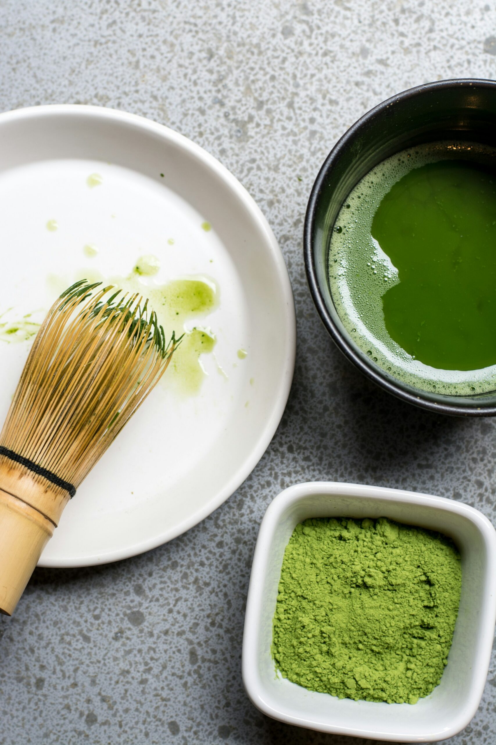 Vue en plongée d'un fouet Chasen sur une assiette blanche, à côté d'un bol de Matcha mousseux et d'une coupelle de poudre de thé vert de haute qualité. Ce kit est une parfaite idée cadeau thé pour initier les amis au rituel du Matcha ou pour compléter les accessoires d'un amateur.