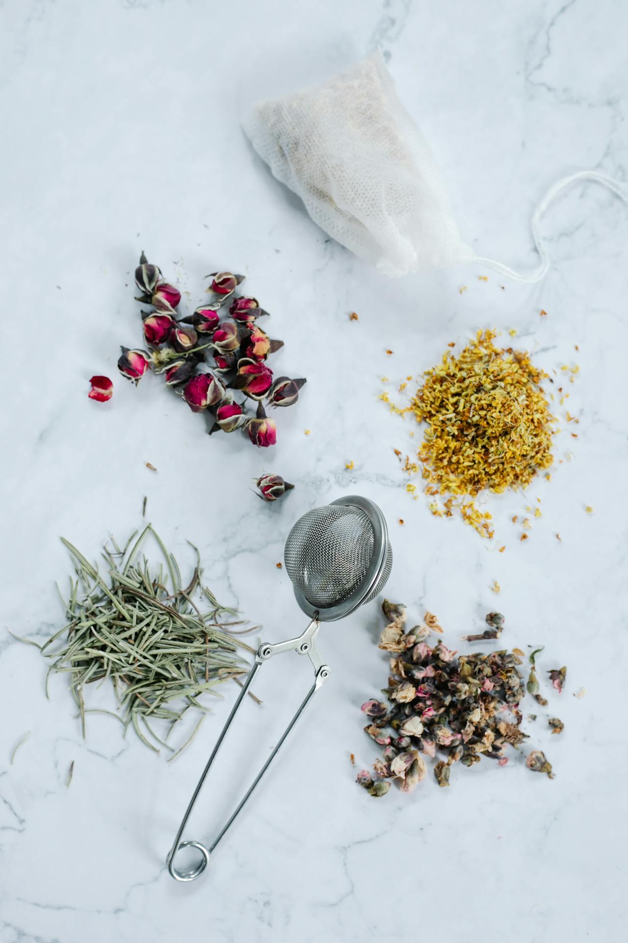 Une boule ou passoire à thé en métal est posée sur un fond de marbre blanc, entourée de divers ingrédients pour infusions : boutons de rose séchés, fleurs d'osmanthus et feuilles de romarin ou de thé blanc. C'est une idée cadeau thé Noël abordable et pratique pour les amateurs de tisanes et de mélanges de fleurs