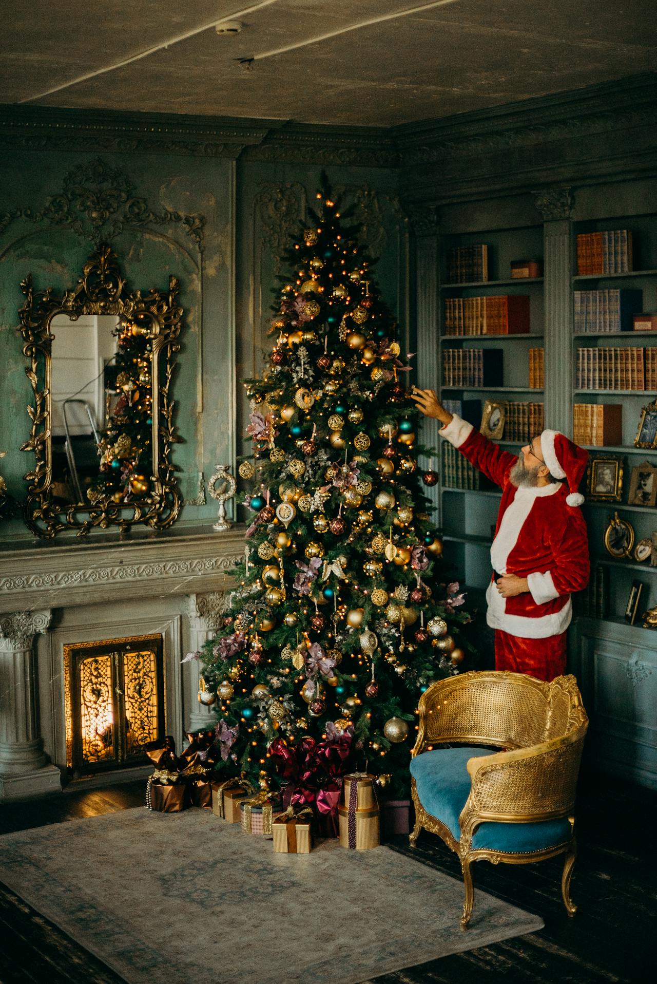 Le Père Noël ajustant une décoration sur un grand sapin de Noël illuminé dans un salon luxueux d'inspiration ancienne avec miroirs dorés et bibliothèque. Cette image illustre l'esprit des fêtes associé à la recette thé de Noël maison.