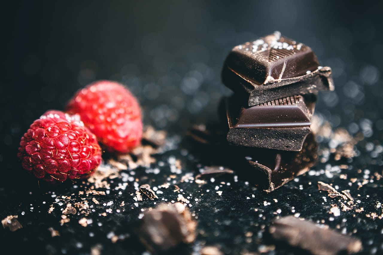 Gros plan sur des carrés de chocolat noir décorés de morceaux de framboise. L'image met en valeur la note acidulée et fruitée, idéale pour un accord thé noir ou un thé vert comme accompagnement.