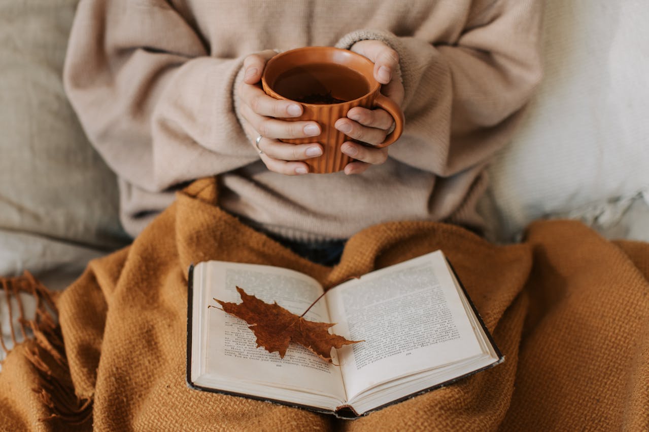 Une personne est assise, tenant une grande tasse de thé chaud à deux mains, sur une couverture douillette. Un livre ouvert est posé sur ses genoux avec une feuille d'automne. C'est l'illustration parfaite d'une idée cadeau thé Noël axée sur le réconfort, la lecture et les moments de bien-être à la maison.
