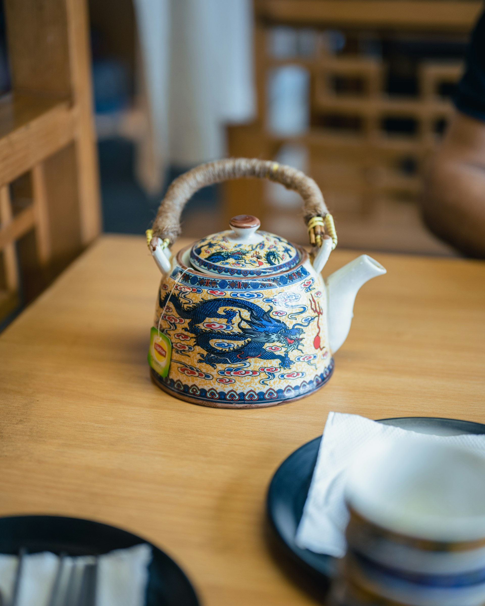Une théière en céramique joliment décorée de motifs de dragon bleu et d'une anse tressée est posée sur une table en bois clair. Une tasse est visible au premier plan, suggérant une dégustation. C'est une excellente idée cadeau thé pour les amateurs de culture chinoise ou les collectionneurs de belles théières.