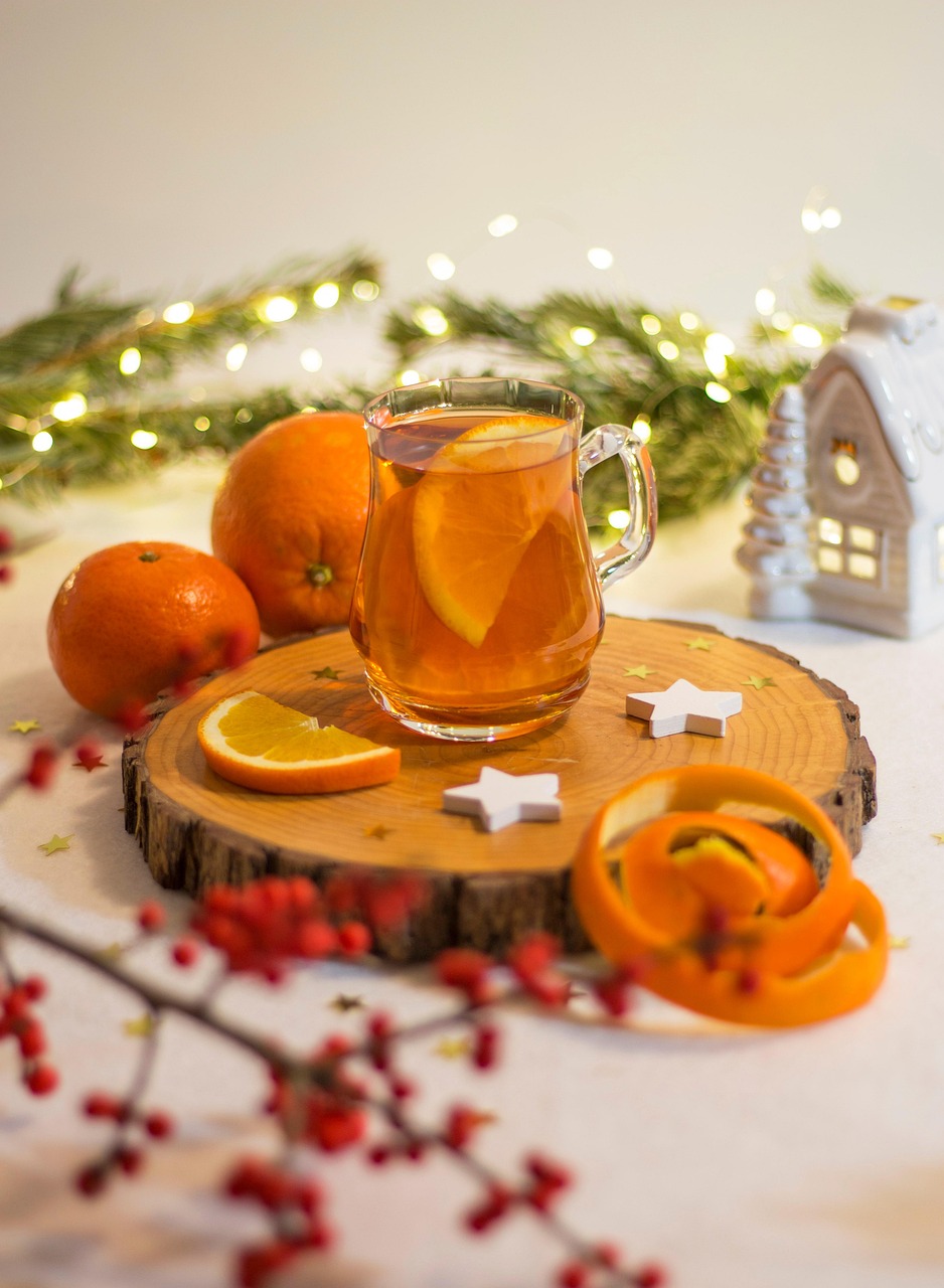 Tasse en verre remplie de thé de Noël aux agrumes avec une tranche d'orange fraîche, posée sur une bûche de bois. Décorations de Noël lumineuses, guirlande, zeste d'orange en spirale et baies rouges en arrière-plan, soulignant le côté fruité de la recette thé de Noël maison.