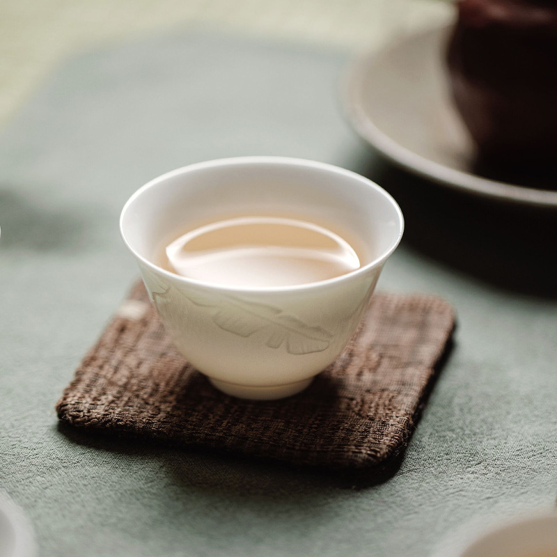 Une tasse en porcelaine blanche délicate remplie de thé blanc vieilli, reconnu comme le meilleur thé pour la digestion, posée sur un dessous de verre texturé.