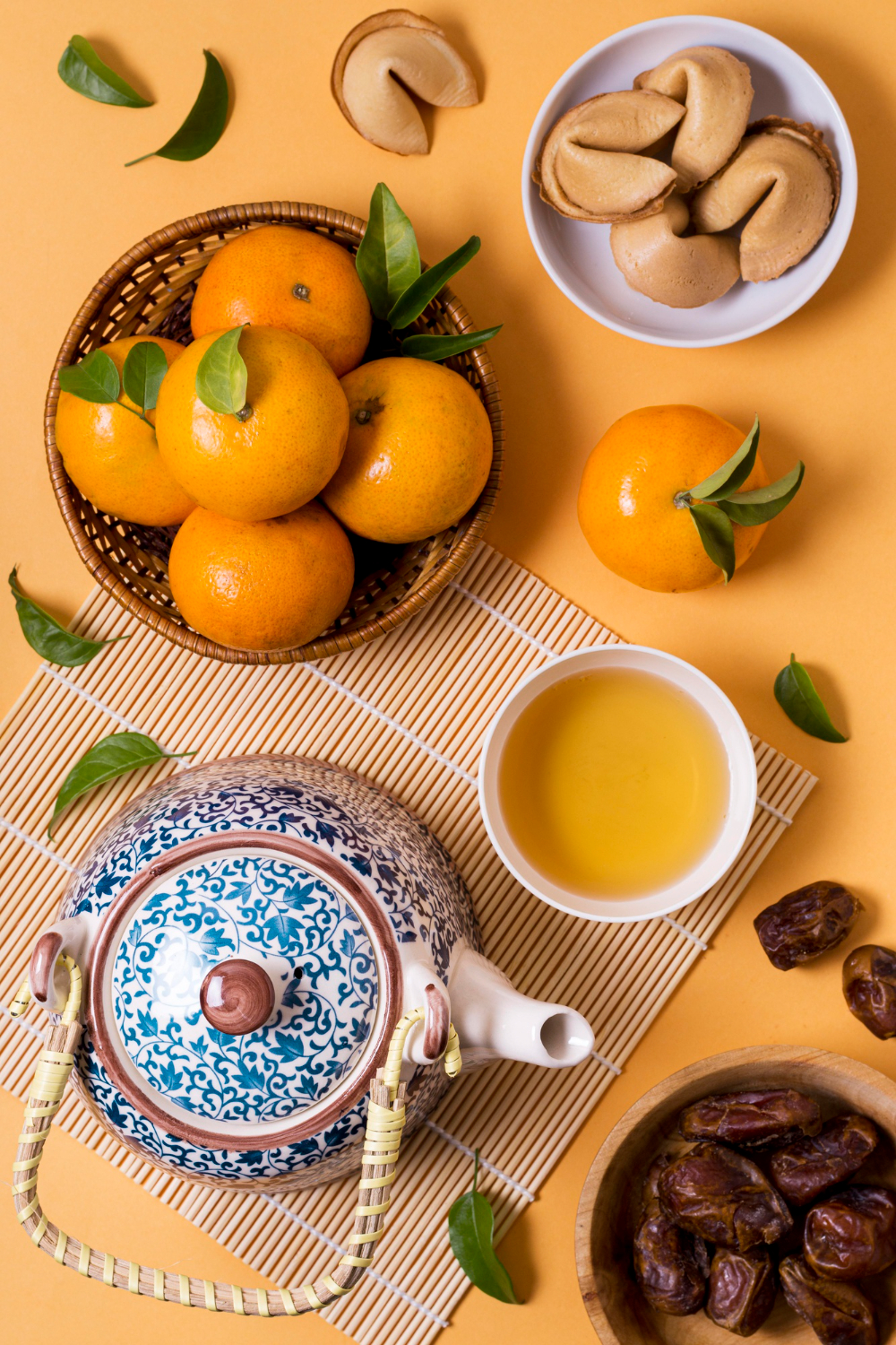 Une vue de dessus d'un service à thé traditionnel avec une théière en porcelaine bleue et blanche et une tasse d'infusion dorée, illustrant l'importance du thé chinois et chance pour Nouvel An chinois. La scène est agrémentée de mandarines fraîches dans un panier, de dattes et de biscuits de fortune, symbolisant la prospérité et les festivités.