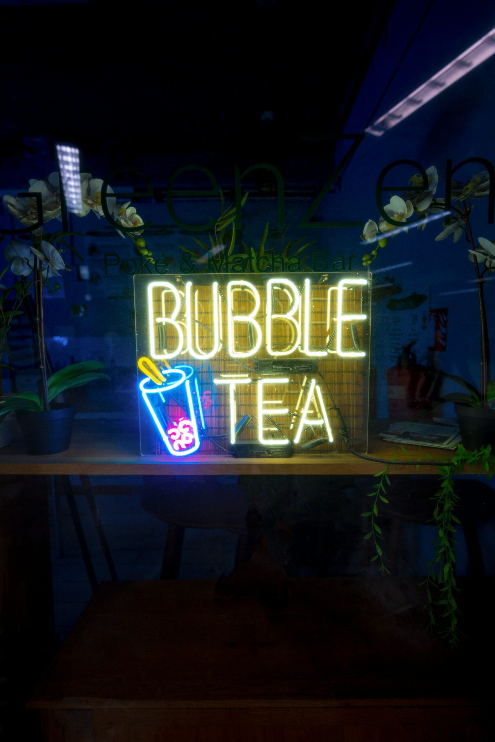 Un néon lumineux jaune et blanc affichant les mots "BUBBLE TEA" avec une icône de gobelet bleue et rose, installé derrière une vitrine décorée d'orchidées blanches. Ce visuel évoque l'ambiance nocturne et moderne où l'on peut déguster un bubble tea à Shanghai dans les quartiers branchés.