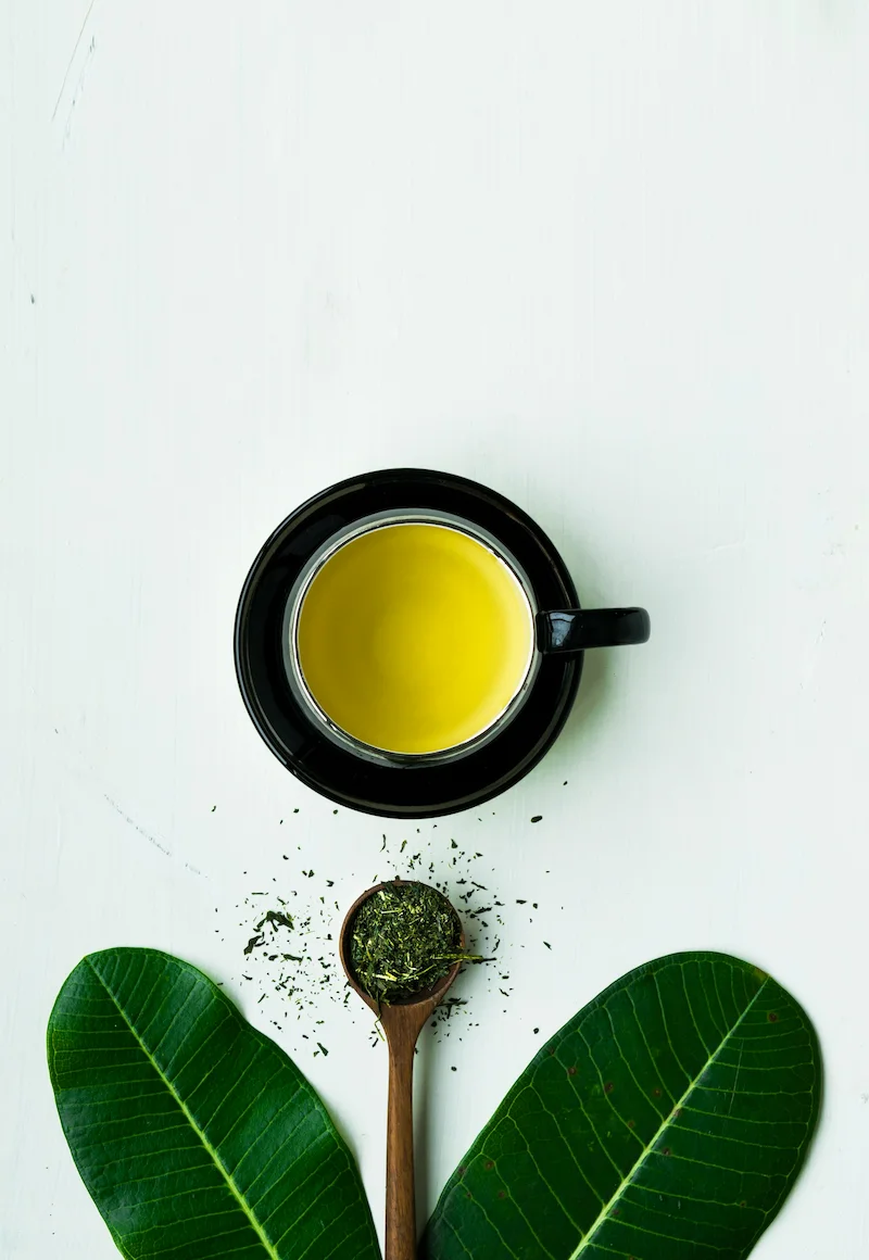 Une tasse de thé vert, souvent considéré comme le meilleur thé pour la digestion, présentée sur une table en bois blanc avec des feuilles de thé en vrac, populaire en France pour ses vertus apaisantes après les repas.