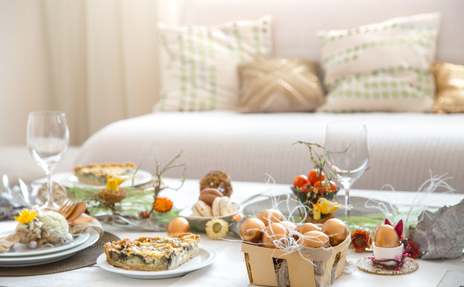 Table de brunch de Pâques élégante avec des parts de quiche aux légumes, un panier d'œufs dorés décoratifs, des macarons et une décoration florale printanière dans un salon lumineux.