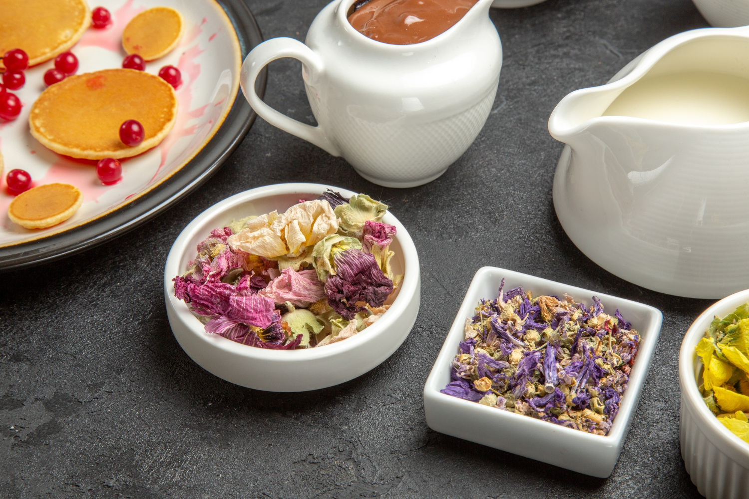 Mise en scène d'un nid botanique d'infusion avec des bols blancs remplis de fleurs séchées colorées (mauve, camomille), accompagnés de petits pots de lait et de chocolat fondu pour un brunch de Pâques.