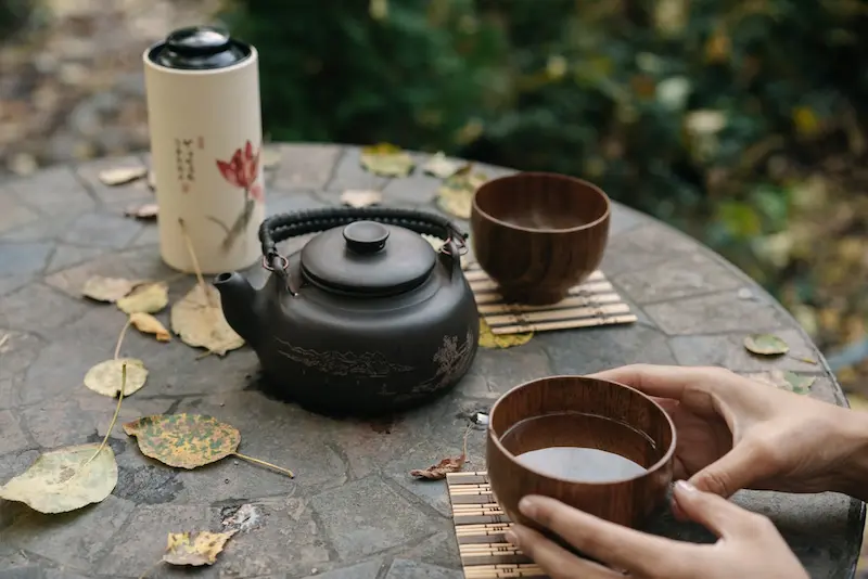Une personne tient un bol en bois rempli de thé entre ses mains sur une table de jardin parsemée de feuilles d'automne. À côté sont posés une théière noire traditionnelle, un second bol et une boîte à thé (thermos) illustrée, invitant à boire de l'eau chaude pour un moment de détente en extérieur.