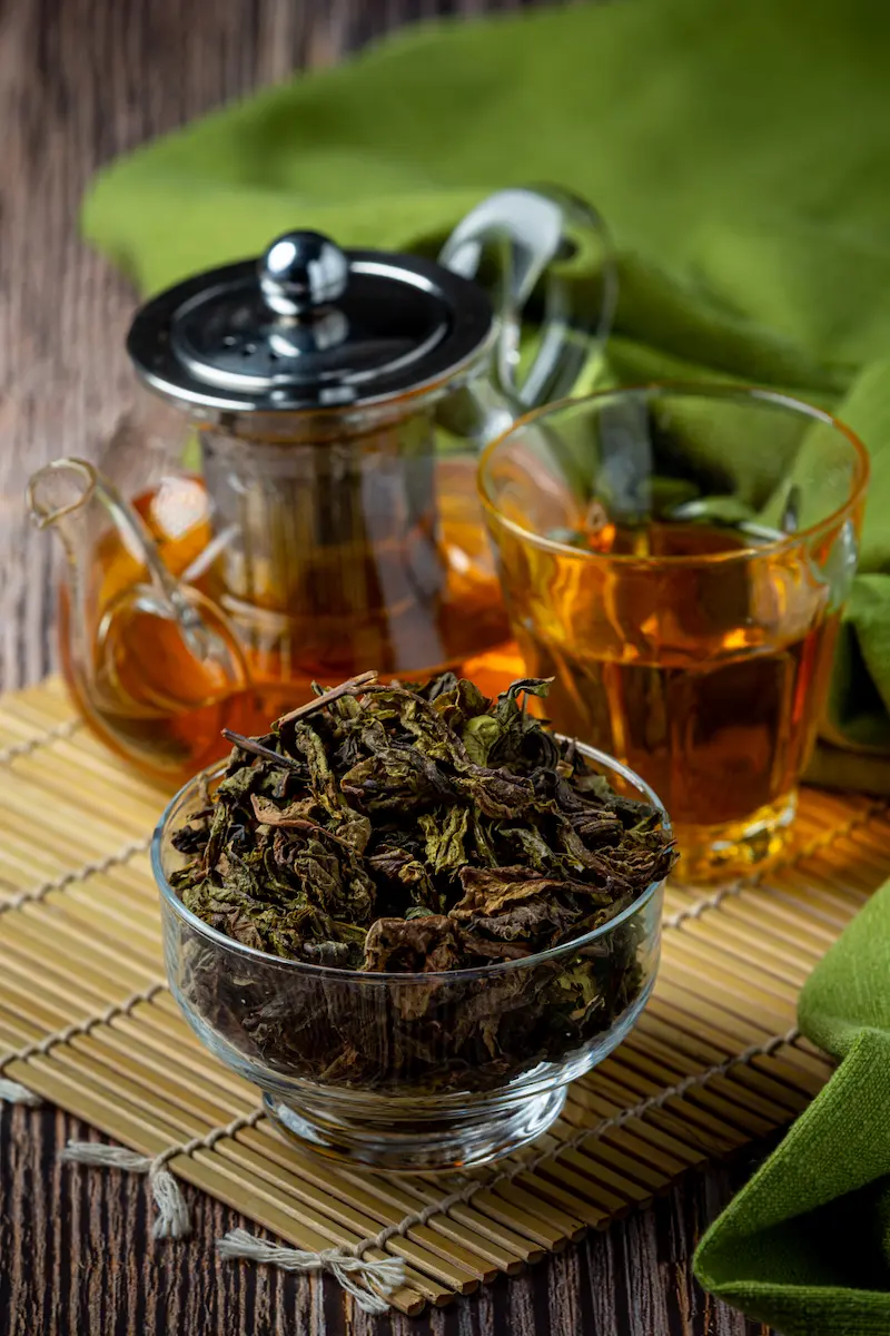Gros plan sur des feuilles de thé Oolong séchées dans un bol en verre, illustrant la richesse du thé chinois et chance pour Nouvel An chinois. En arrière-plan, une théière en verre transparente et un verre rempli d'une infusion ambrée sont posés sur un set de table en bambou, évoquant une dégustation traditionnelle et raffinée.