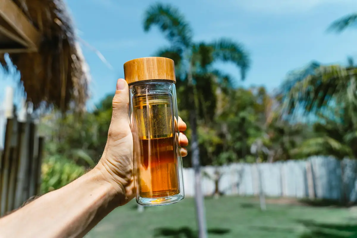 Gros plan sur une main tenant une bouteille infuseur en verre (thermos) remplie de thé ambré sous un ciel bleu et des palmiers. Le visuel illustre pourquoi les Chinois boivent de l'eau chaude partout, afin de profiter des bienfaits de l'eau chaude et d'une infusion saine même en vacances ou en extérieur.