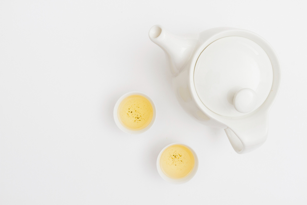 Vue de dessus d'une théière en céramique blanche et de deux petites tasses de thé blanc infusé sur fond blanc. Le thé blanc est idéal pour comprendre la différence théine et caféine grâce à sa libération d'énergie très douce.