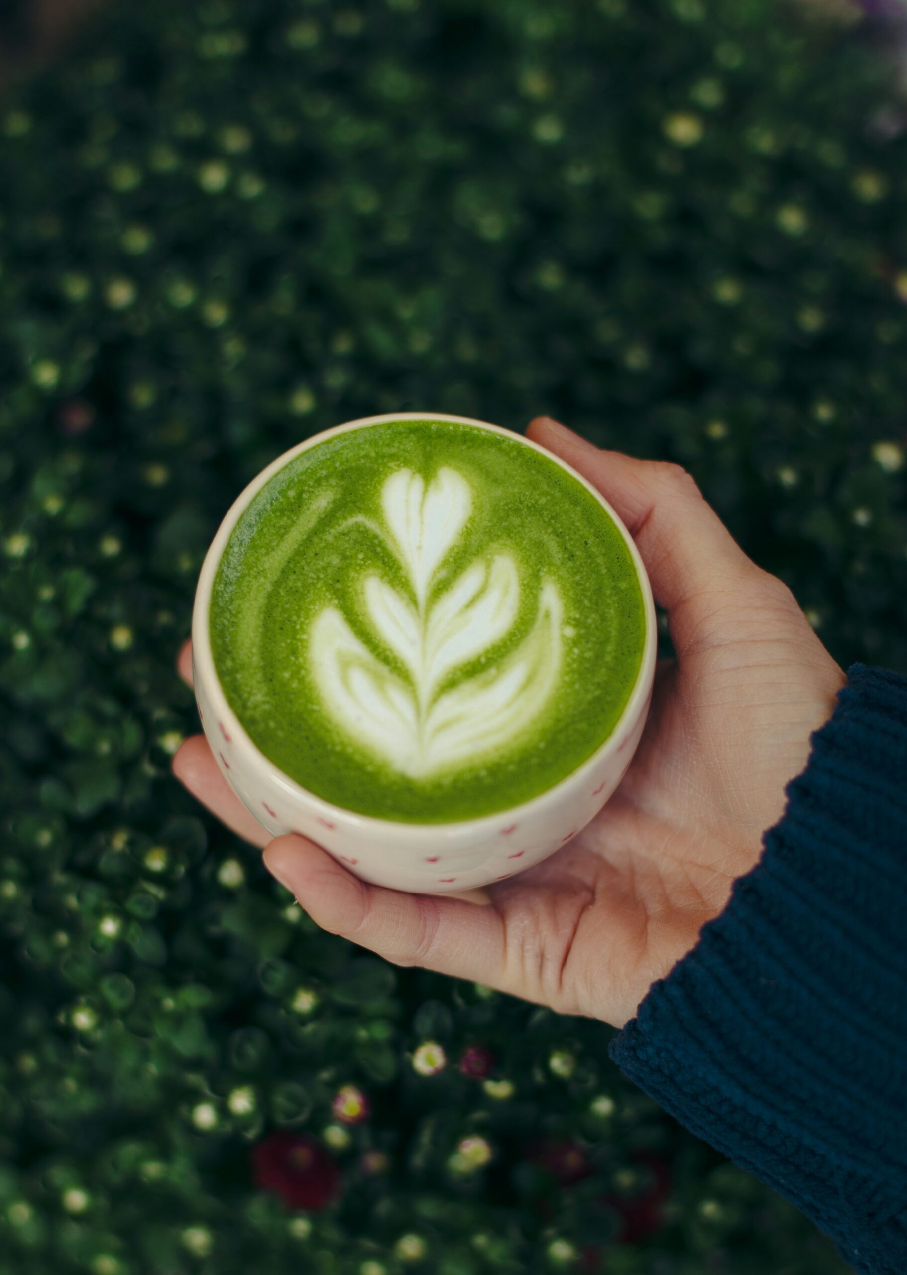 Main tenant une tasse de matcha latte onctueux avec un latte art en forme de fleur, sur un fond de feuillage vert. Le matcha est une excellente alternative au café pour une énergie stable.