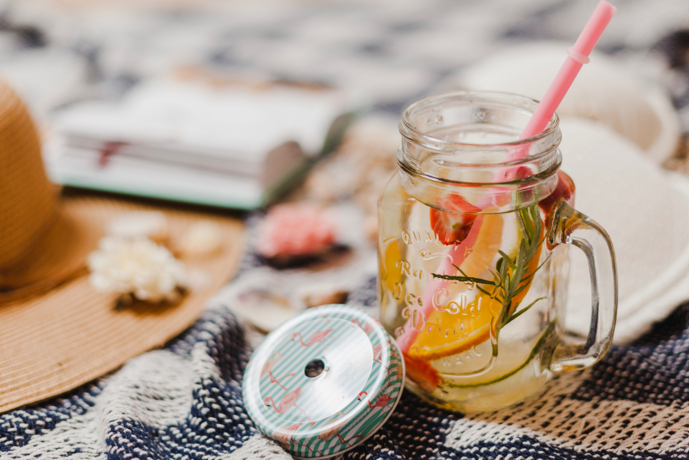 Au premier plan, un thé glacé infusé avec des fruits frais (agrumes, baies et menthe) présenté dans une Mason jar au couvercle métallique. À l’arrière-plan, une plage ensoleillée avec sable clair et mer bleue. Une boisson rafraîchissante obtenue par infusion à froid, parfaite pour les journées chaudes. Exemple idéal de Thé infusion à froid à déguster en vacances.