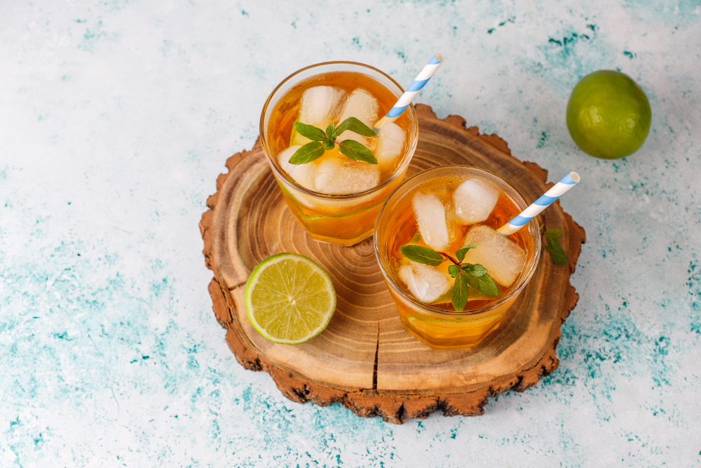 Deux verres de thé glacé au citron vert et à la menthe, posés sur un rondin de bois. Chaque verre est muni d'une paille. Une boisson fraîche obtenue par infusion à froid, mettant en valeur les arômes naturels des agrumes et de la menthe. Parfait pour illustrer un Thé infusion à froid partagé à deux.