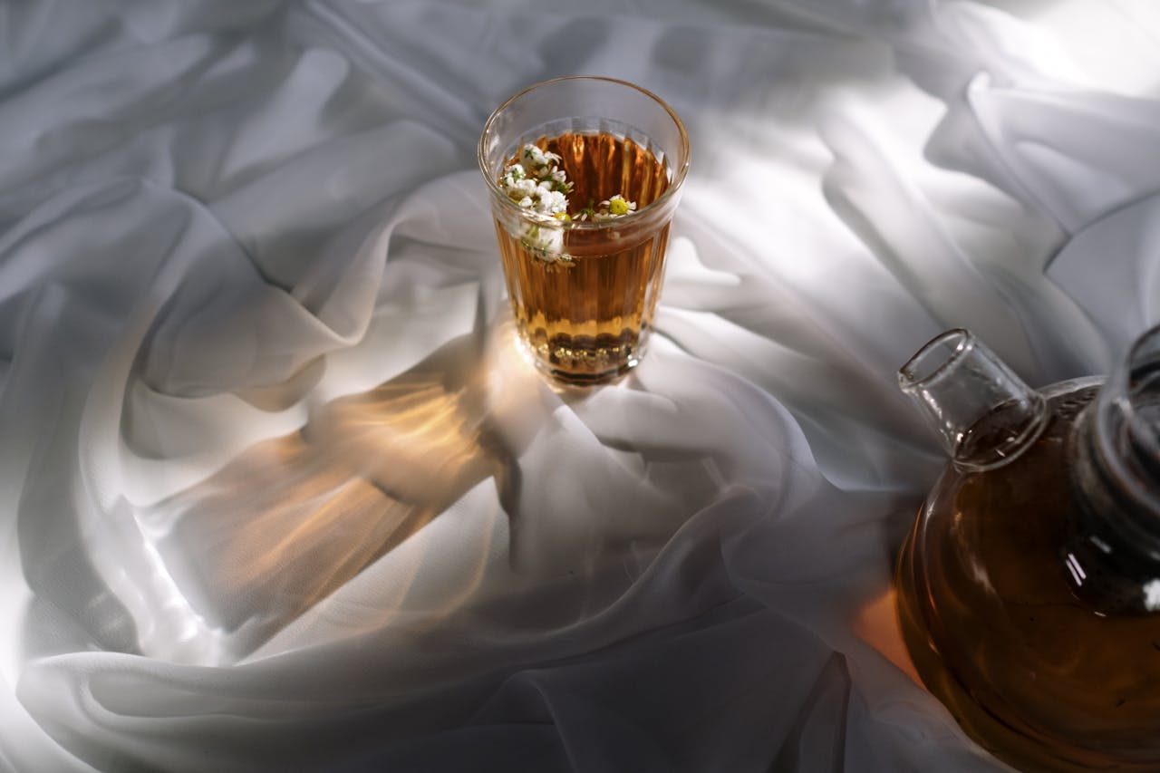 Théière en verre et verre d'infusion chinoise pour dormir aux fleurs blanches, posés sur des draps blancs soyeux pour une infusion sans théine soir apaisante.