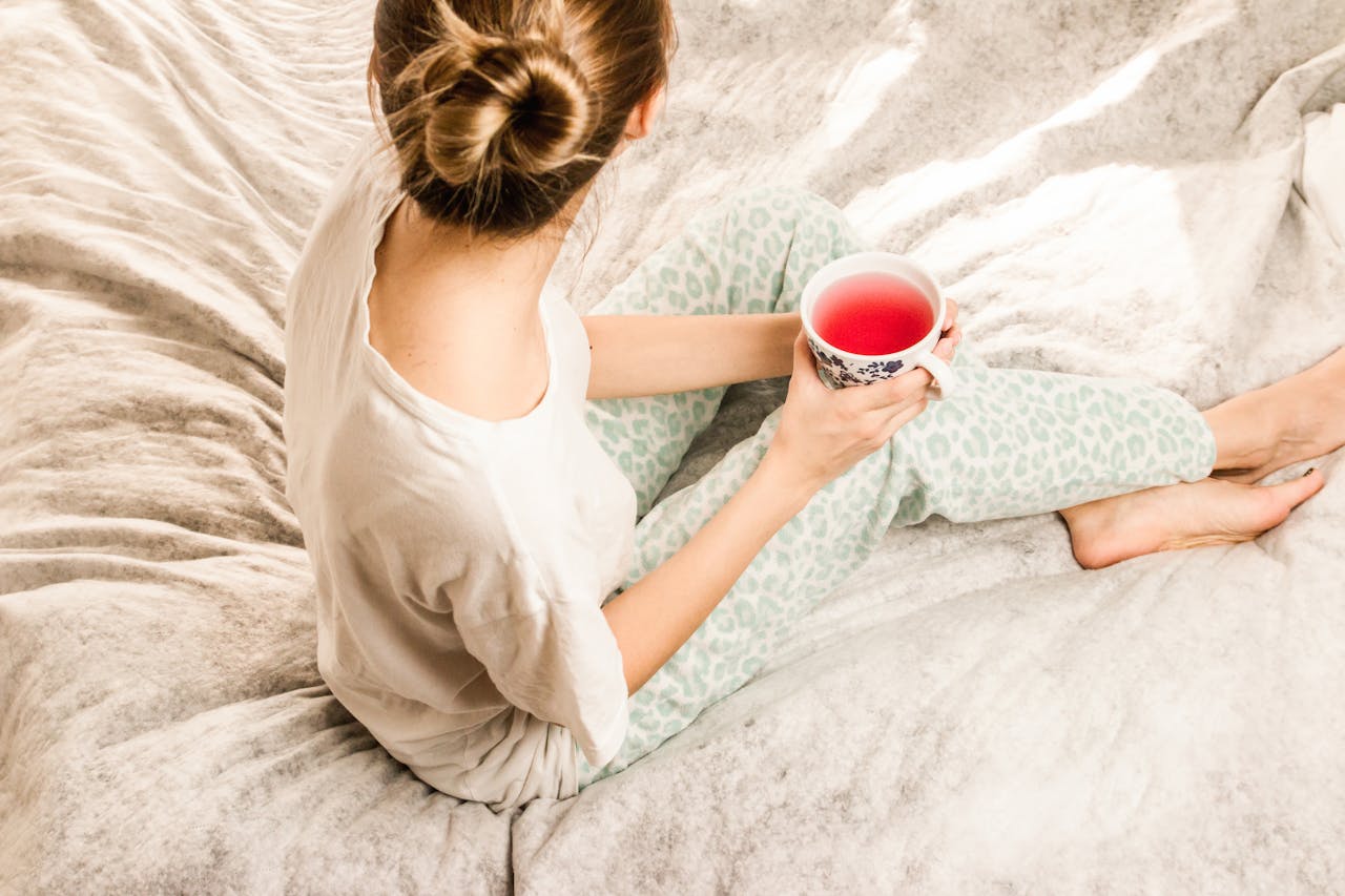 Vue de dessus d'une femme en pyjama sur un lit, tenant une tasse d'infusion à la rose. Cette image illustre une infusion chinoise pour dormir, parfaite comme infusion sans théine le soir dans le cadre d'un rituel soir sans théine.