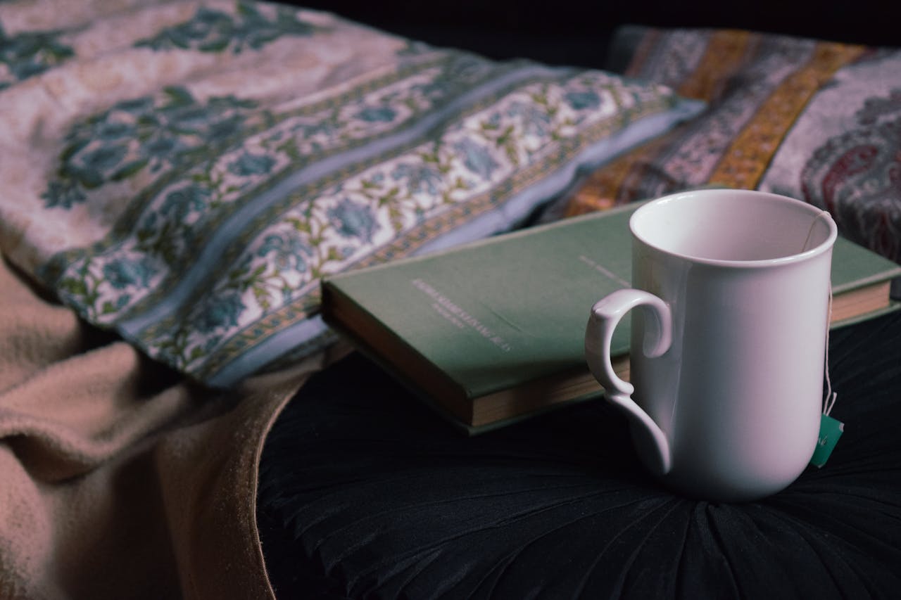 Une tasse fumante posée sur un livre près d'un lit douillet, illustrant une infusion sans théine soir pour un moment de lecture relaxant avant de dormir.