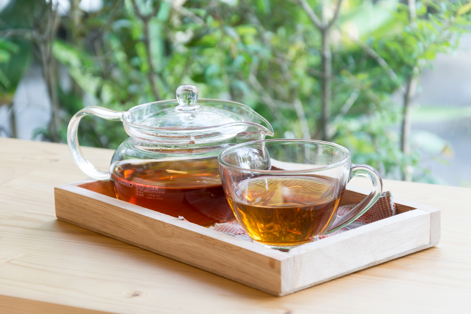 Théière et tasse en verre contenant un thé vert clair, posées sur une table en bois clair. Une pause douce pour boire du thé en voyage ou adopter un rituel thé nomade